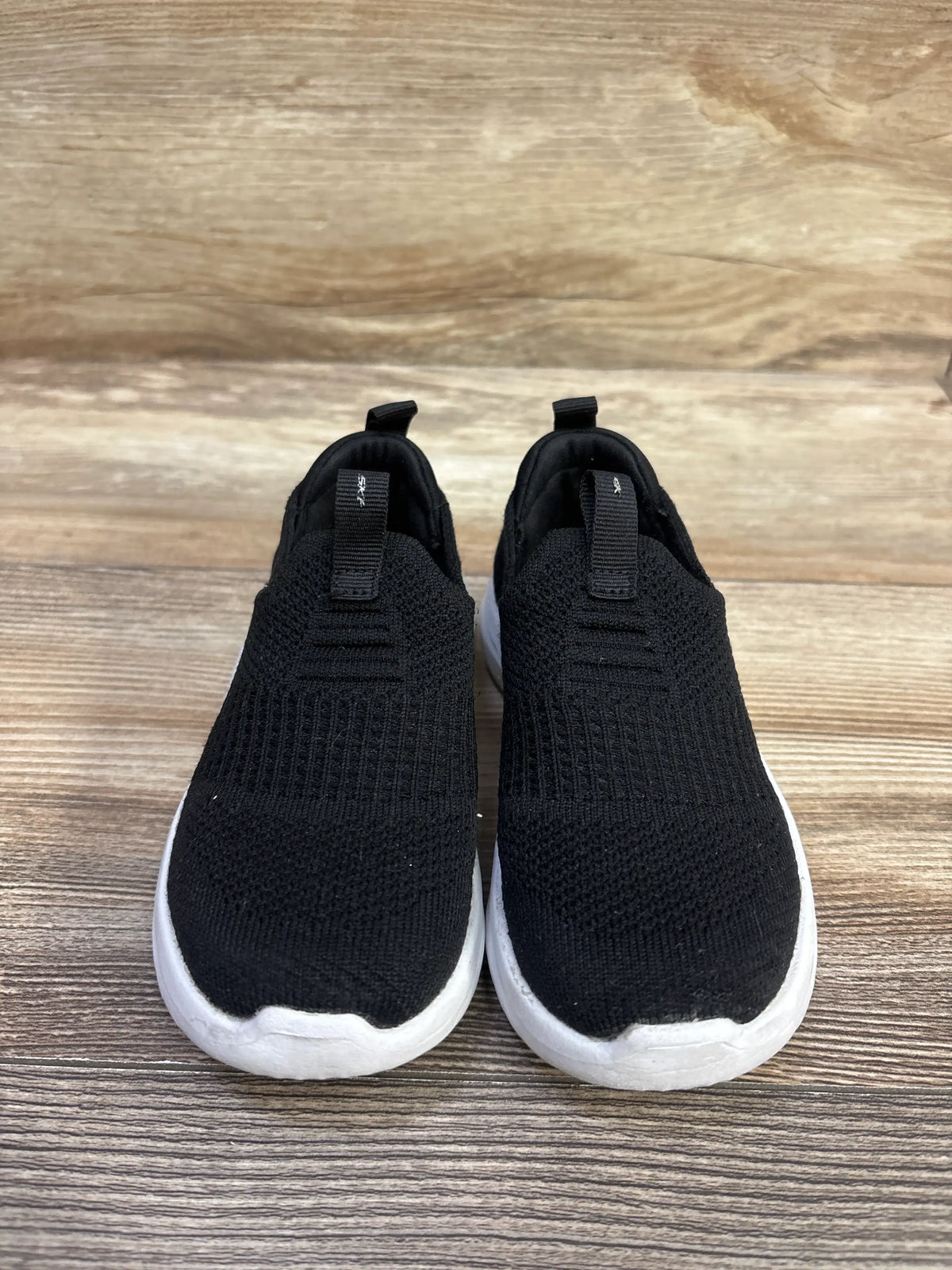 Front view of black Skechers Ultra Flex sock-like sneakers with white soles and heel pull tabs on a wood surface