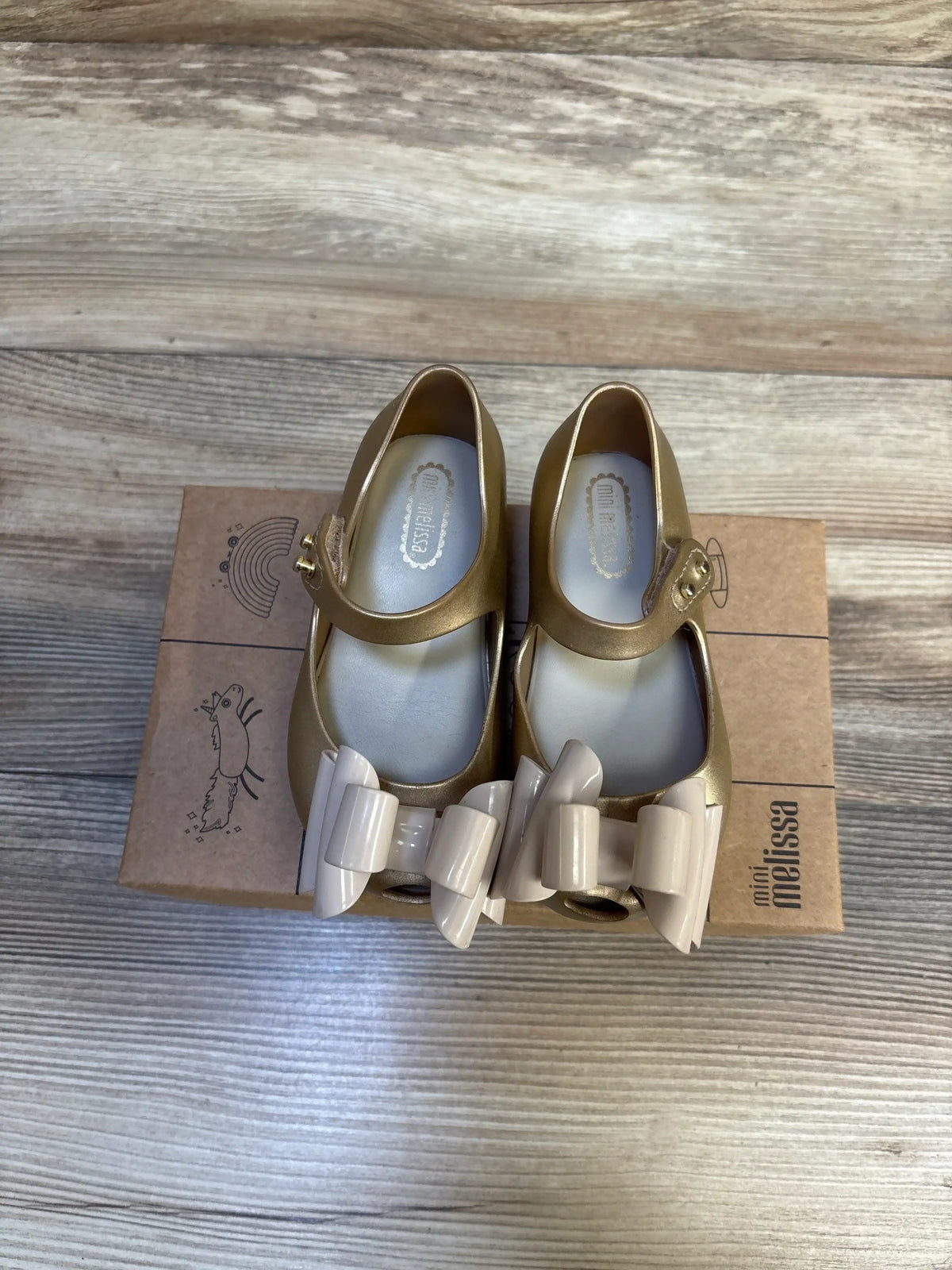 Top view of Mini Melissa Ultragirl Sweet Mary Jane toddler shoes in gold showcasing beige bow accents on a Mini Melissa box over wood floor