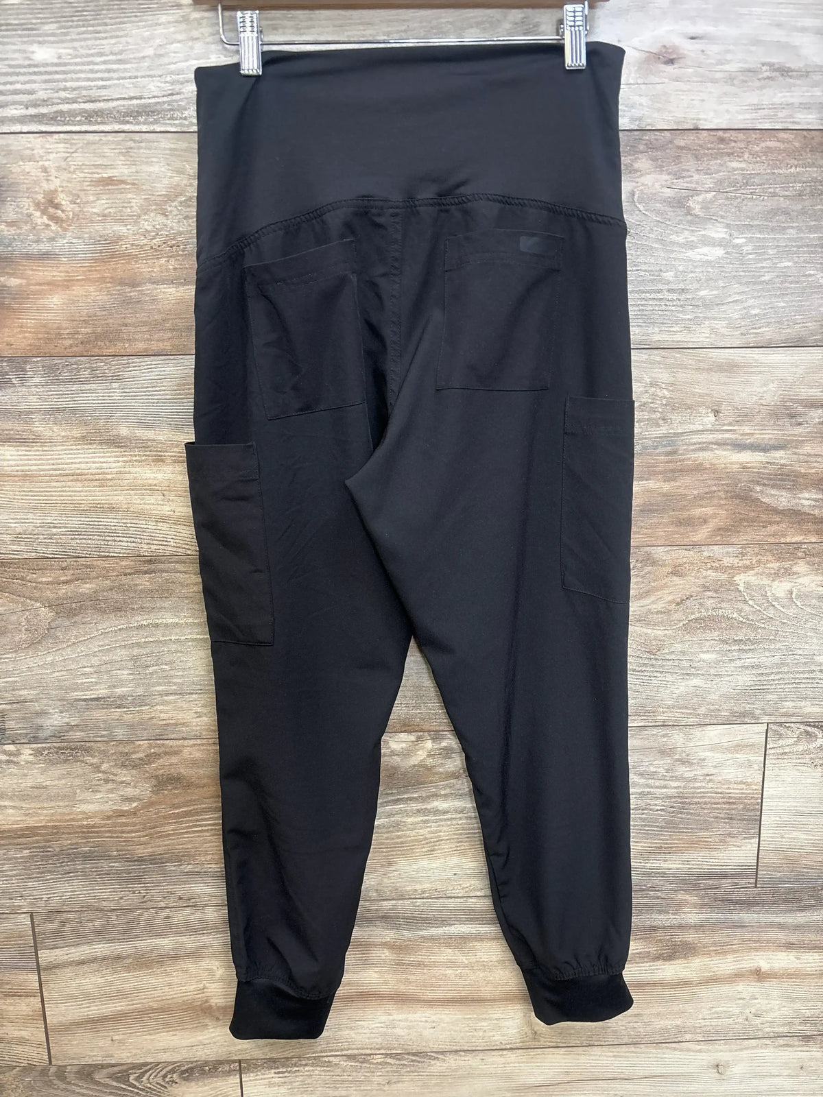 Back view of black maternity scrub joggers showcasing high fold-over waistband, side and back patch pockets, and elastic ankle cuffs on wood plank background