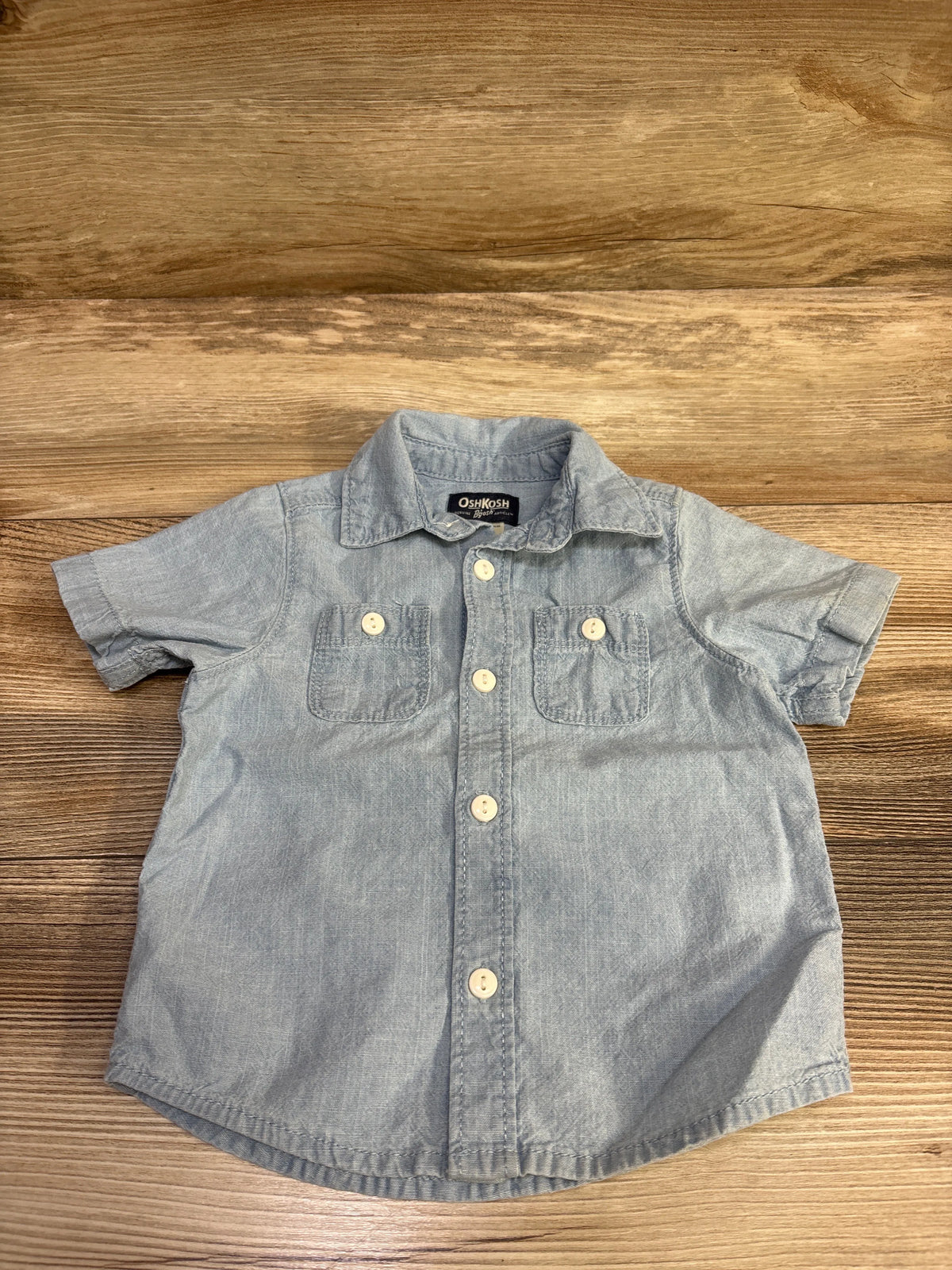 Front view of OshKosh blue chambray button-up shirt with short sleeves, collar, buttons, and two chest pockets on wood grain
