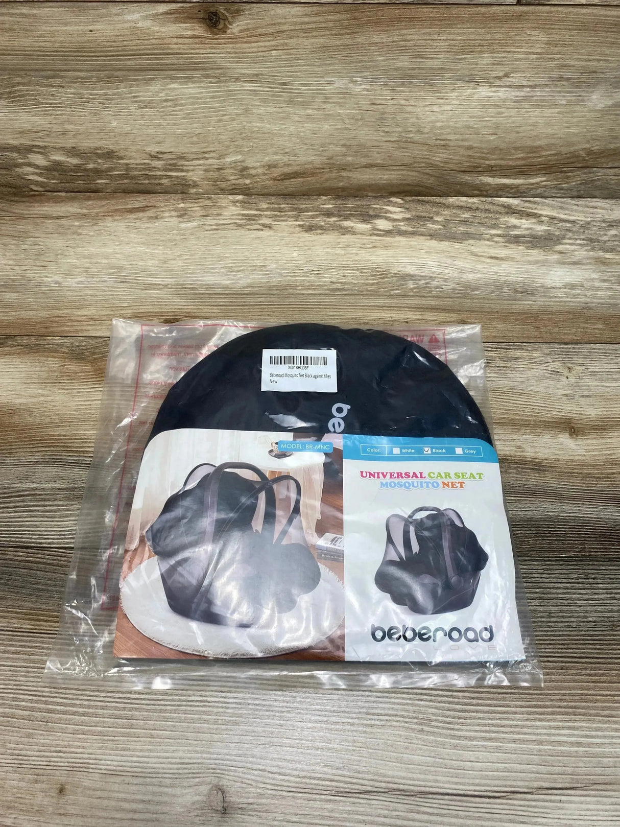 Top down view of Beberoad car seat mosquito net in black, folded in round pouch inside clear plastic packaging on wood surface