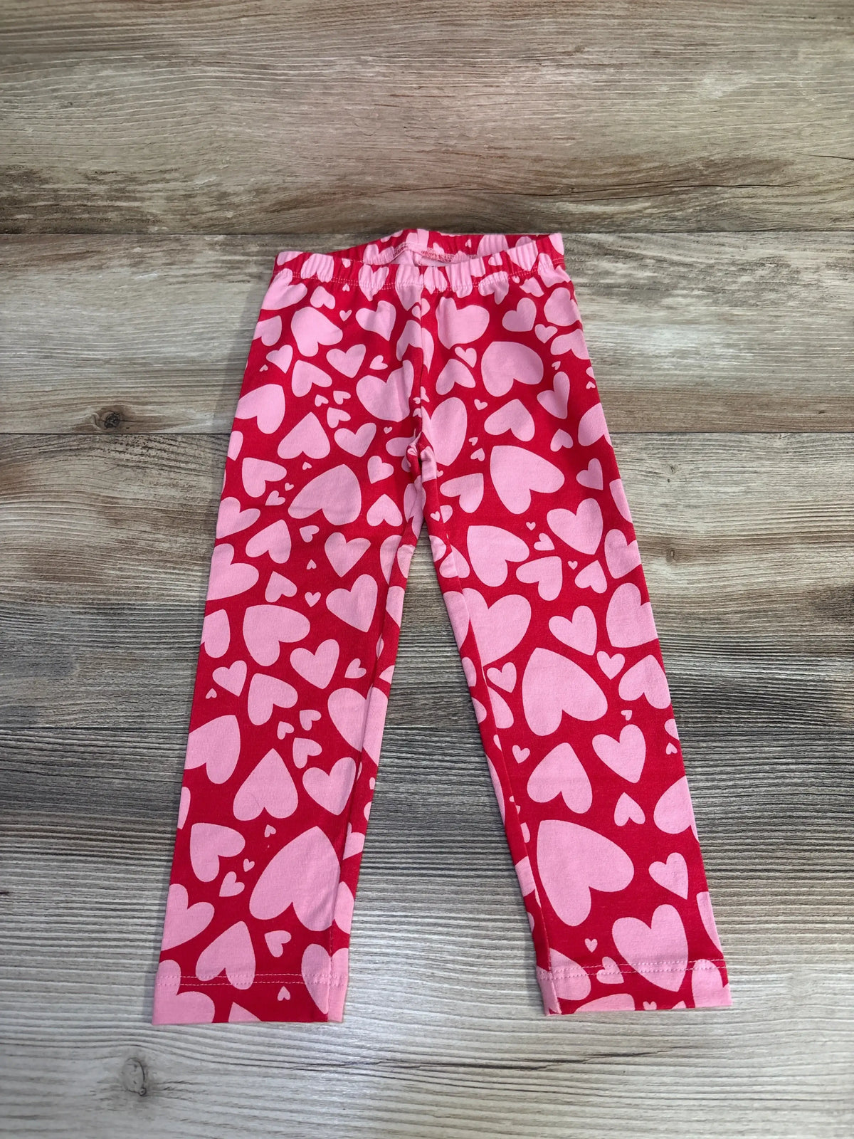 Front view of Old Navy heart leggings in red, size 2T, showcasing a pink heart pattern on a wood-grain background