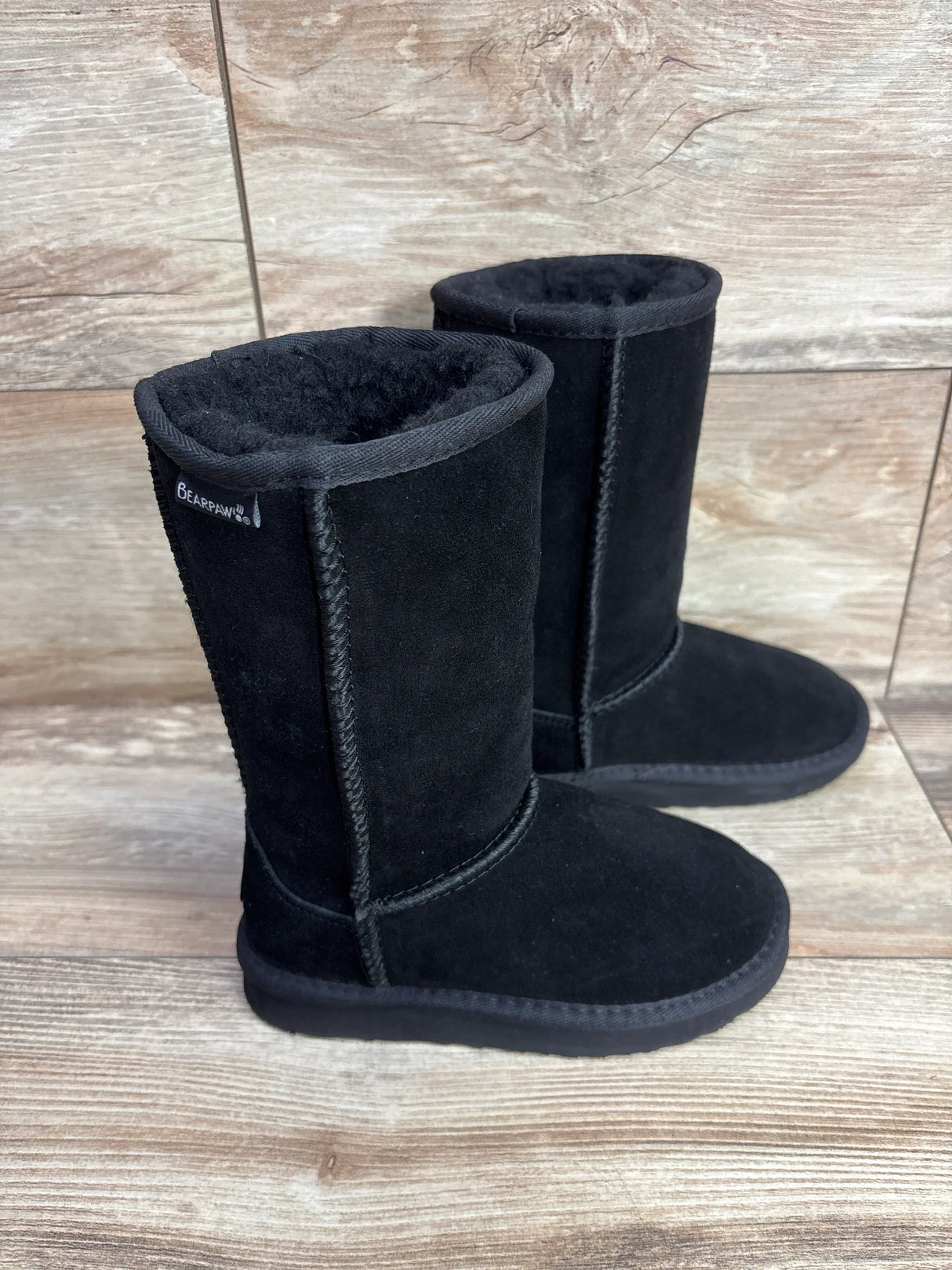 Side view of Bearpaw Kenzie tall wool-lined kids boots in black showing suede exterior and plush lining on wood floor