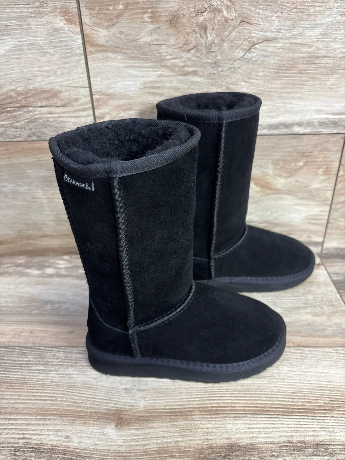 Side view of Bearpaw Kenzie tall wool-lined kids boots in black showing suede exterior and plush lining on wood floor