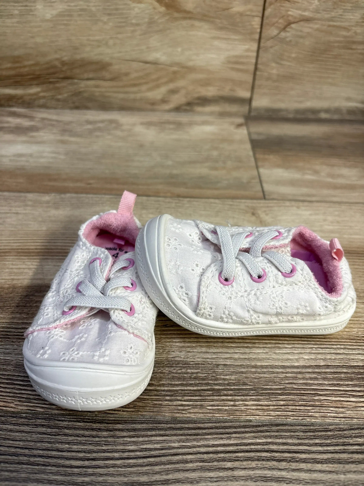 Three-quarter front view of toddler white eyelet sneakers size 4C showcasing pink lining, pink eyelet rings and rubber toe bumper on wood floor