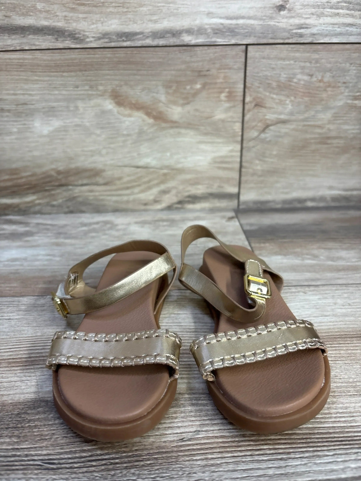 Front view of MIA Peyton toddler sandal in gold with ankle buckle, stitched toe strap, and brown cushioned sole on wood
