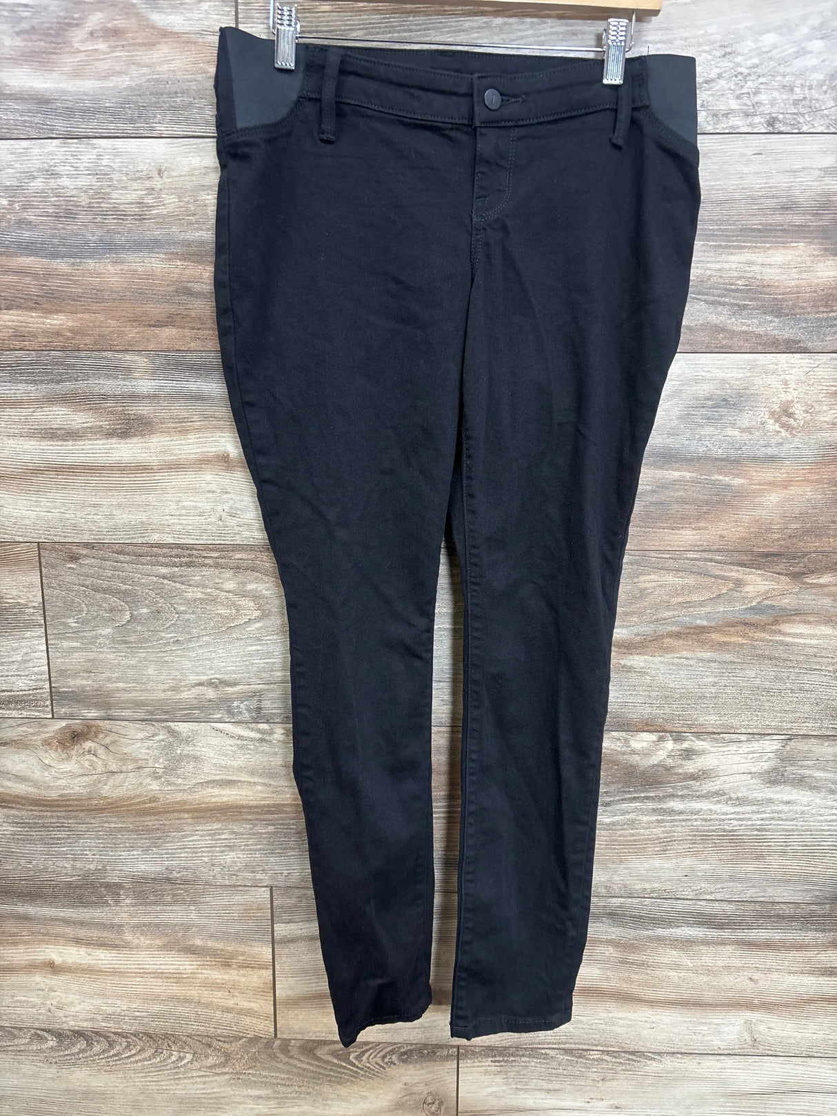 Front view of black Isabel maternity skinny jeans showing elastic side panels and skinny leg, hanging on wood background
