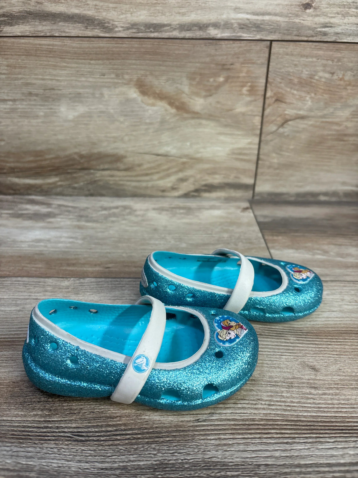 Angled front view of children's glitter blue Crocs Mary Jane flats showing white strap and Frozen character charm on wood floor