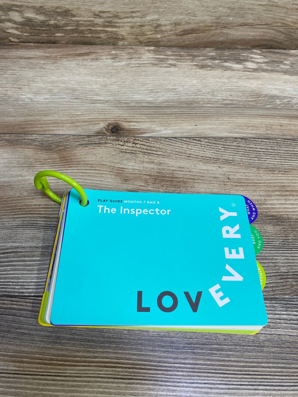 Top down view of Lovevery The Inspector play guide, turquoise cover labeled Months 7 and 8, green ring on wood background