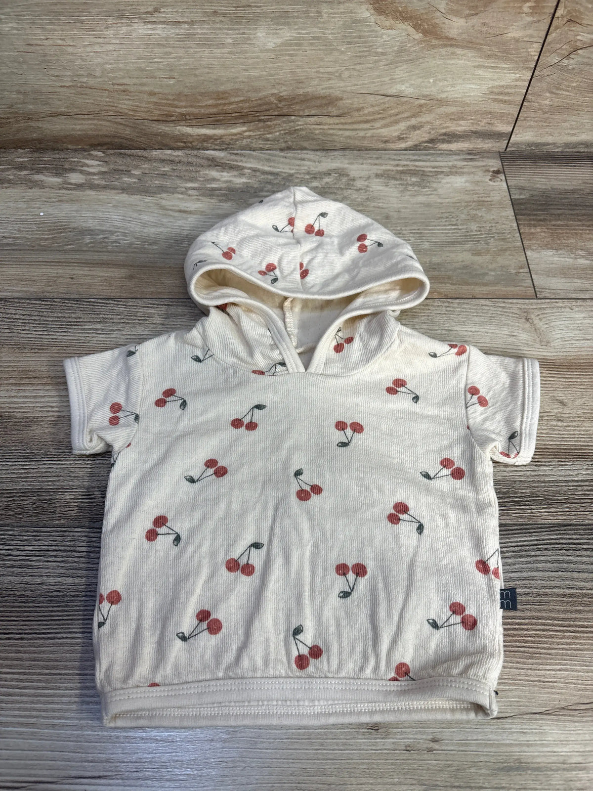 Front view of Modern Moments beige hooded shirt for ages 6-9 months, showcasing cherry print and short sleeves on a wood floor