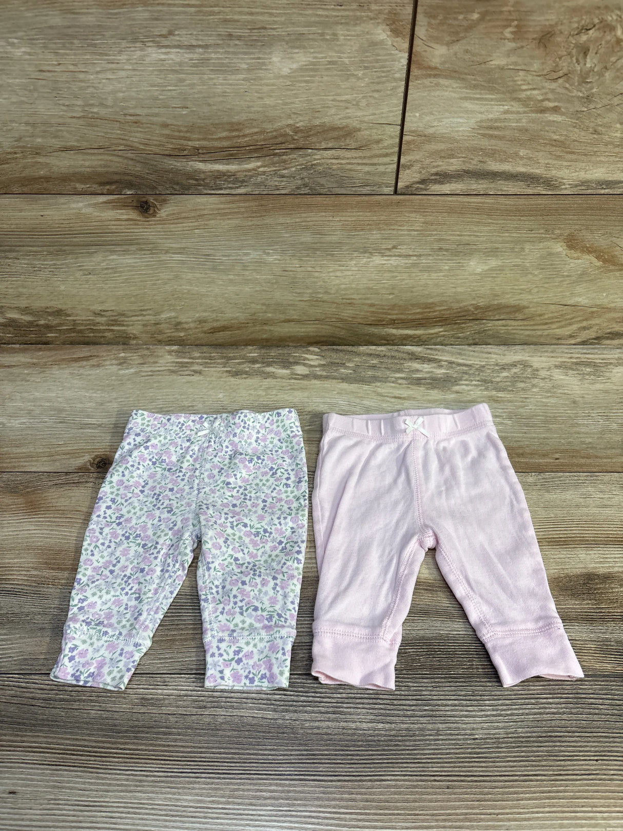 Top-down view of Carter’s 2-pack baby pants (3M) showcasing one lilac floral pant and one solid pale pink pant on wood floor
