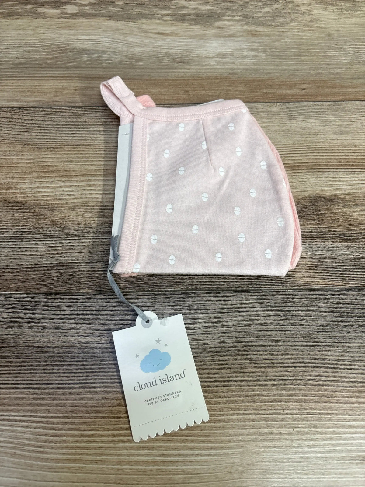 Top-down view of pink Cloud Island newborn bonnet showcasing white dot pattern and brand hang tag on wooden background