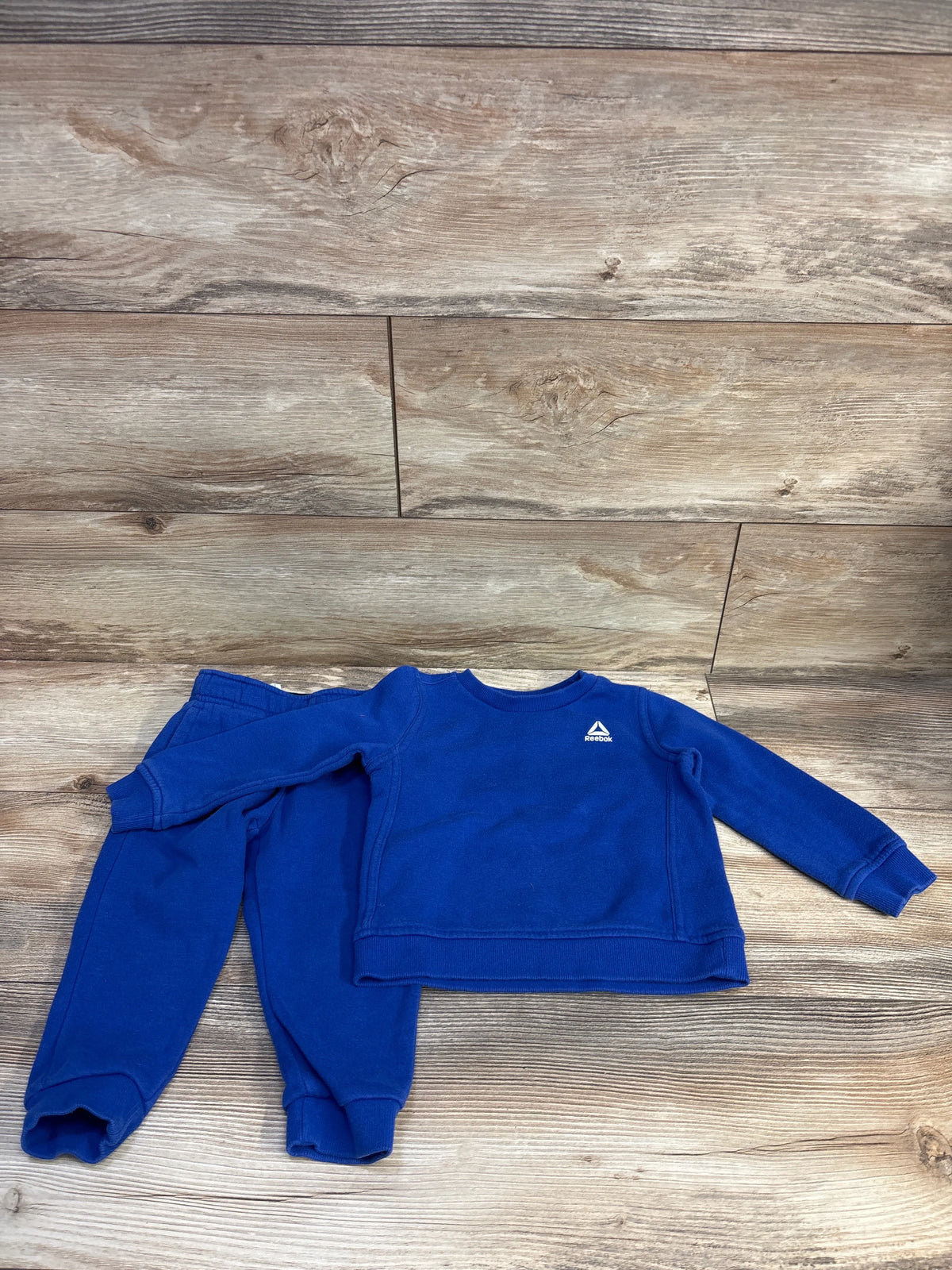 Flat lay of blue toddler Reebok pullover and jogger set showing Reebok logo on chest and elastic cuffs on wood background