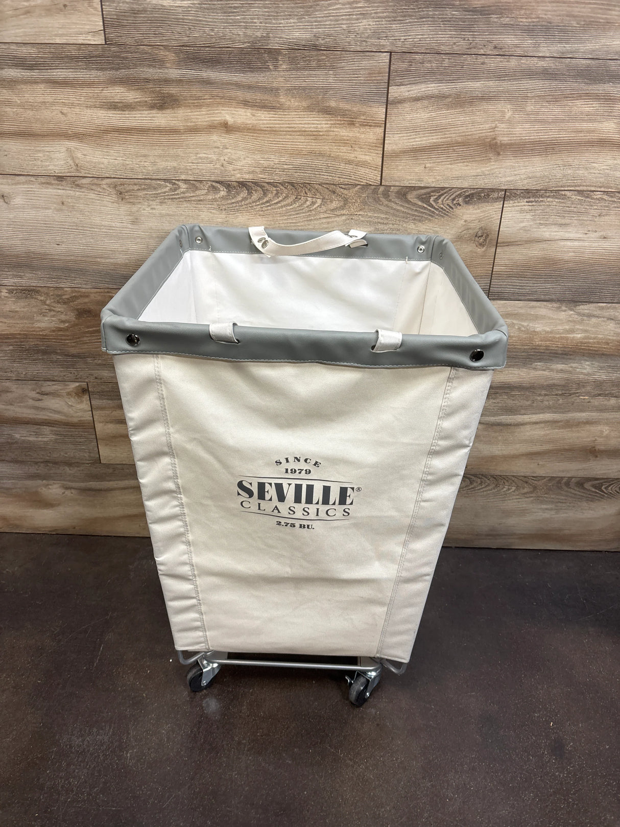 Front view of Seville Classics canvas laundry hamper with gray trim and caster wheels against wood-plank wall