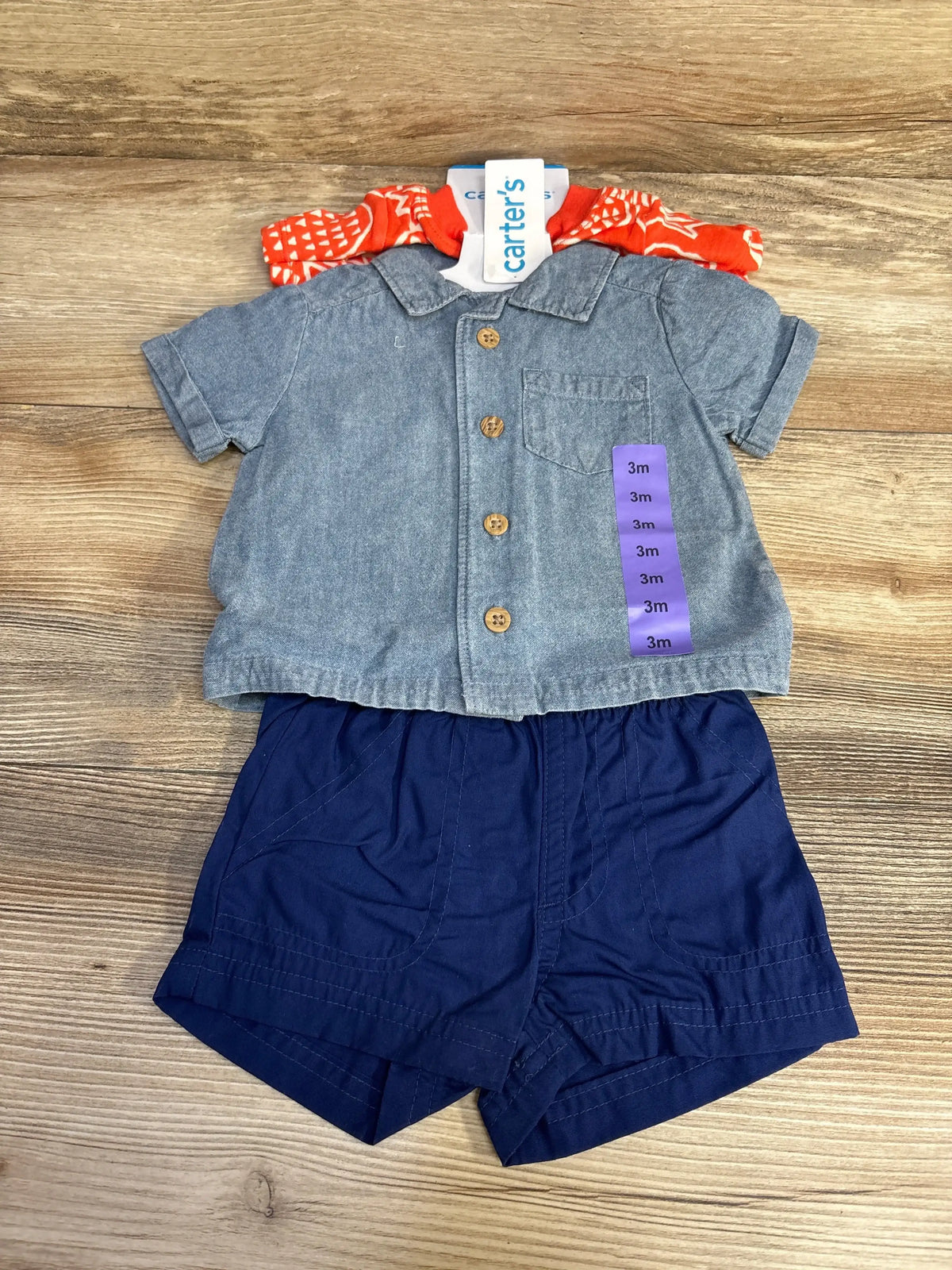 Flat lay of Carter’s 3-piece baby set: chambray button-up, navy shorts, orange onesie; size 3m stickers on wood