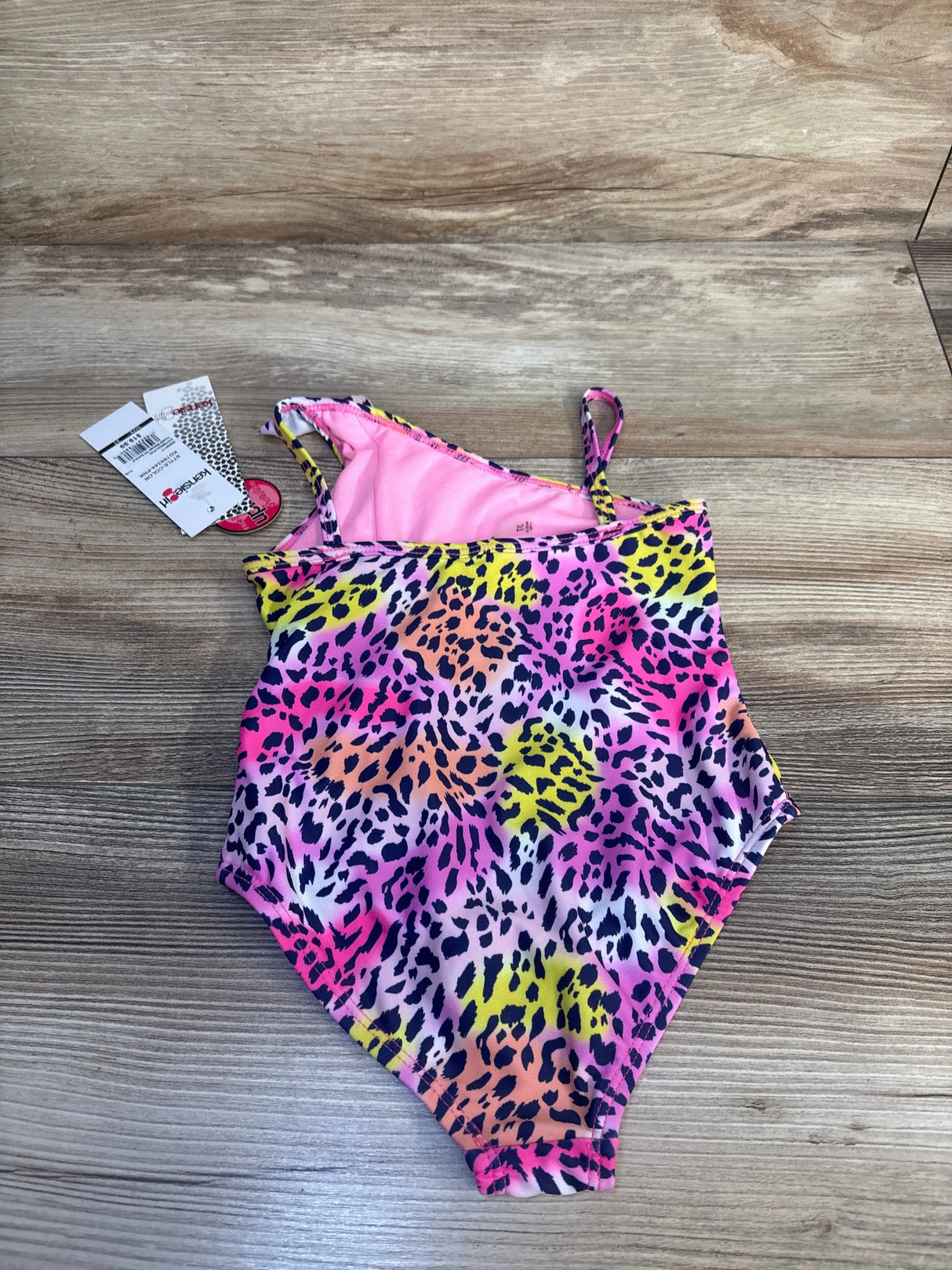 Girls' one-piece swimsuit with colorful leopard print, pink lining, and tags, on wood background