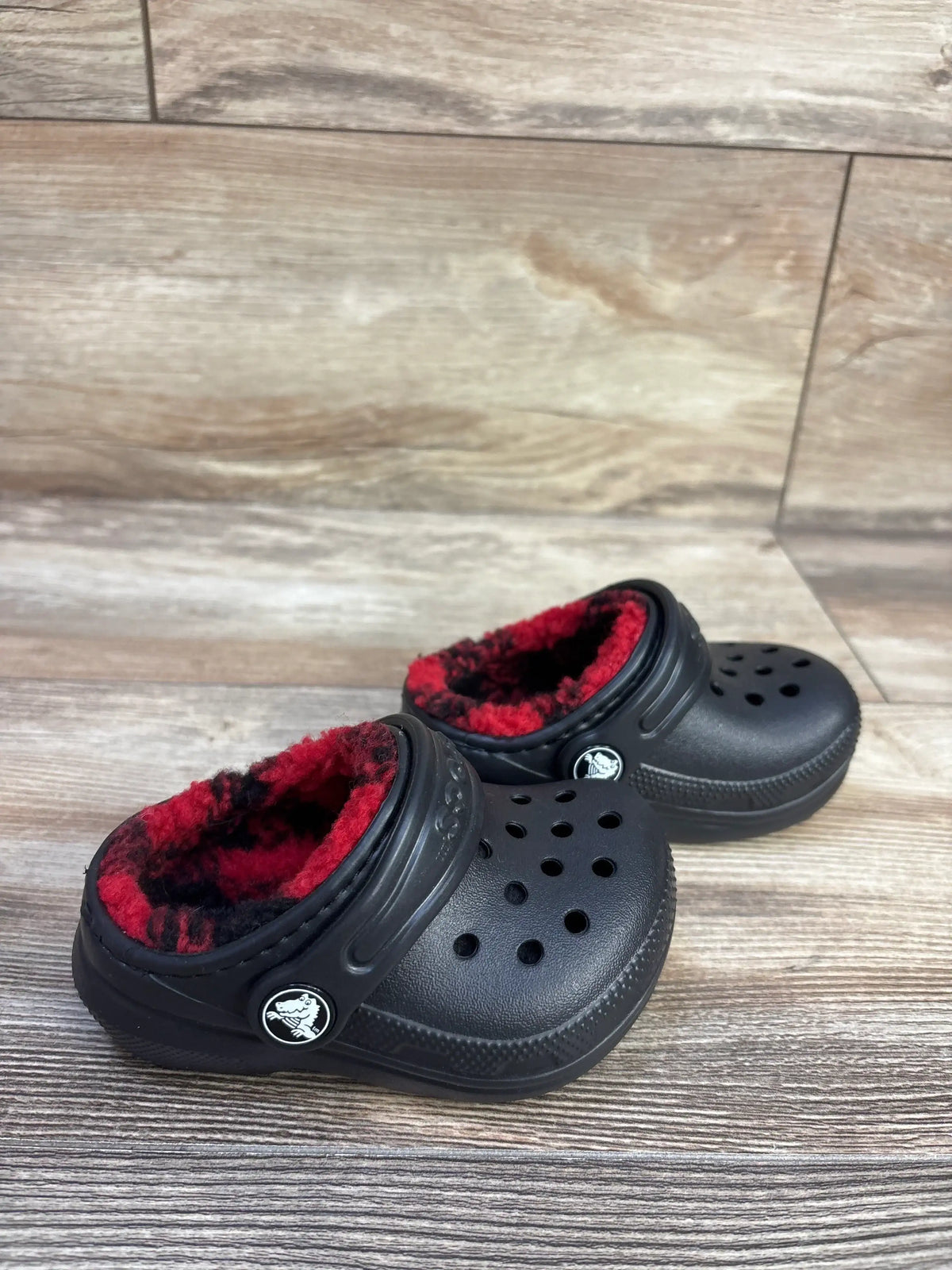 Angled front view of black Crocs Classic lined clogs showing red and black buffalo check fleece lining on wood floor background