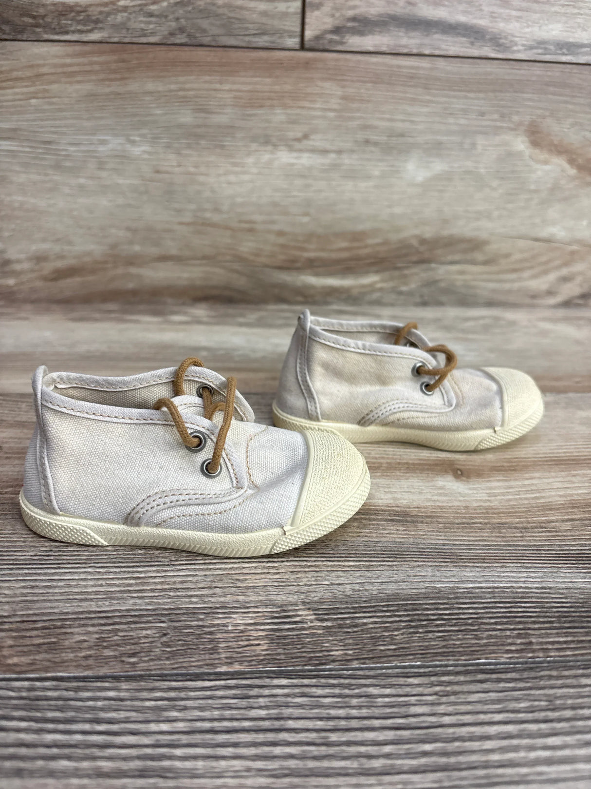 Pair of light beige toddler canvas shoes with brown laces on wooden floor background