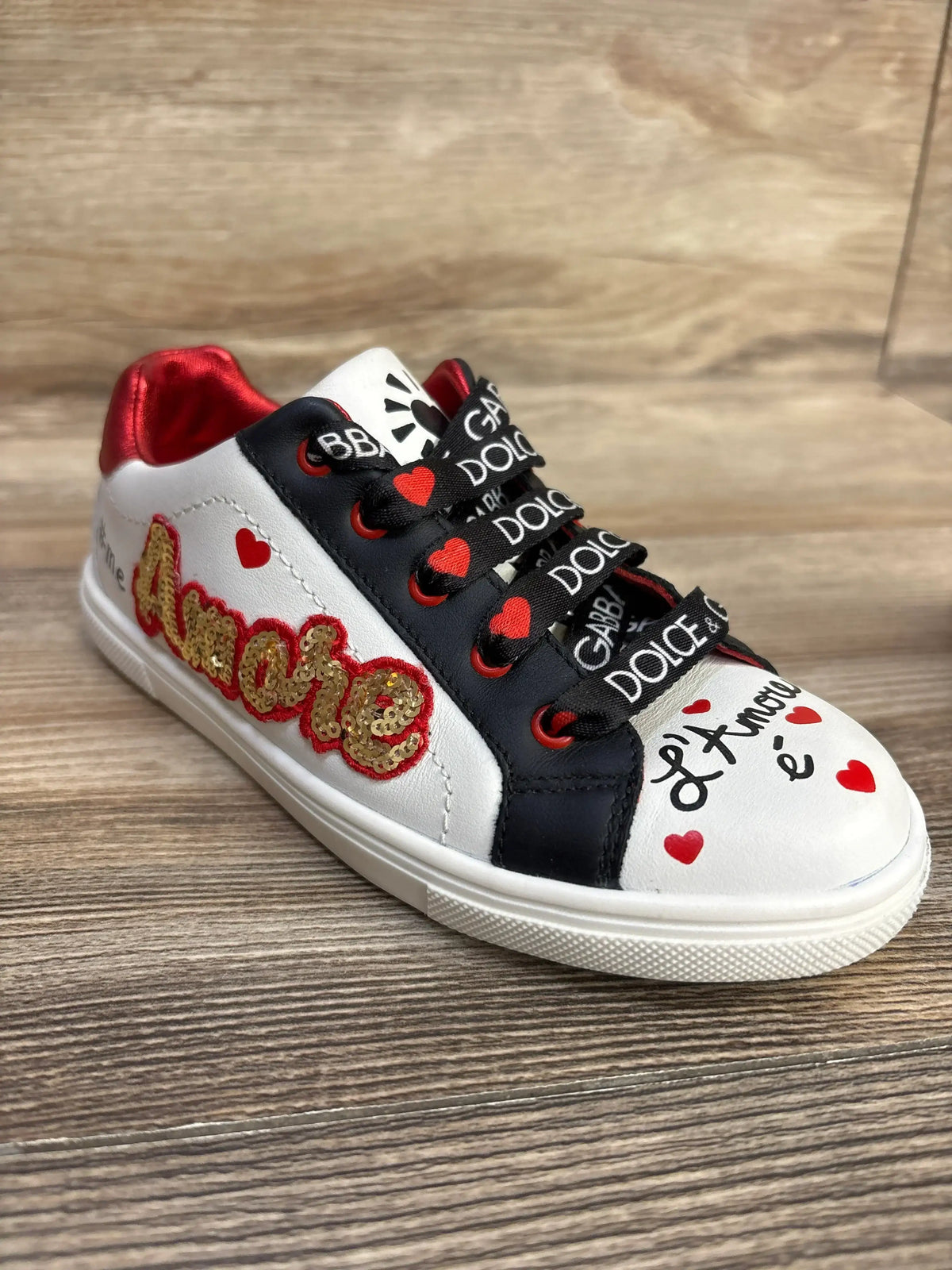 Angled view of Portofino DG Amore sneaker showcasing white calfskin, gold Amore applique, red hearts, and branded laces on a wood background