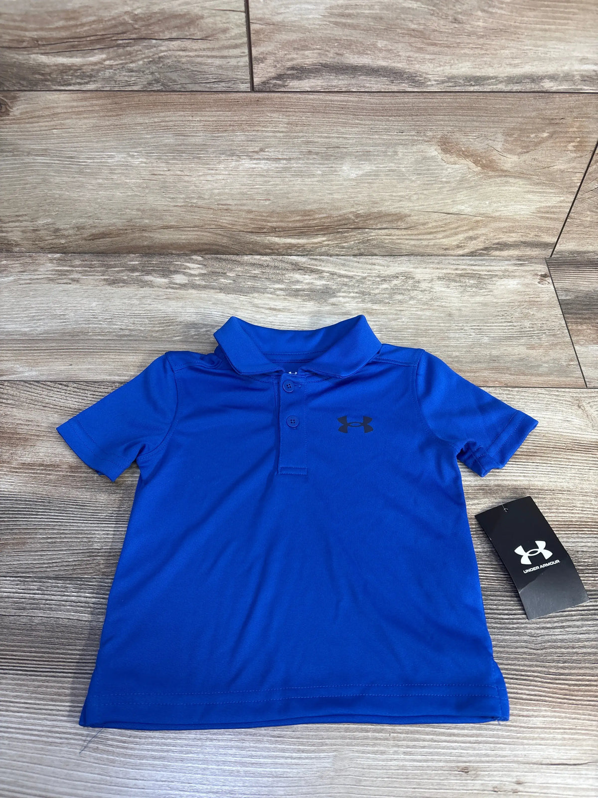 Front view of blue Under Armour polo shirt showcasing collared two-button design and logo on a wood-grain background
