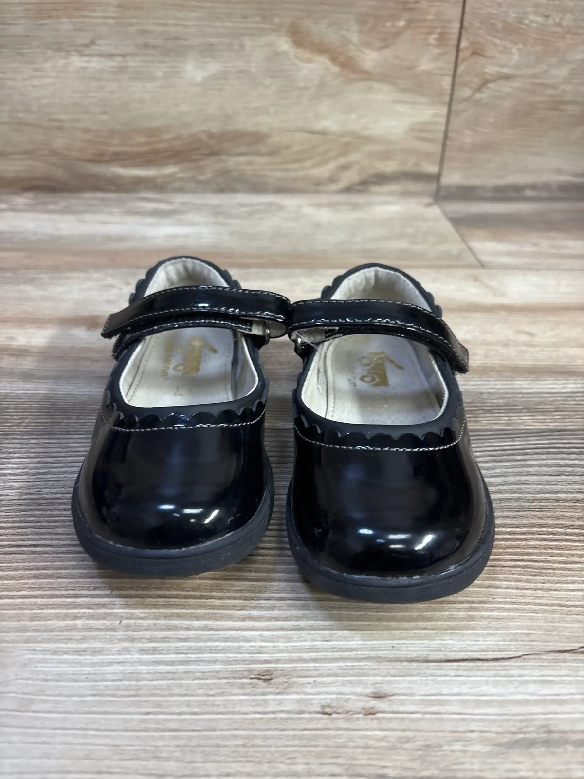 Front view of See Kai Run Mary Jane toddler shoes in black patent leather with scalloped trim and Velcro strap on wood floor