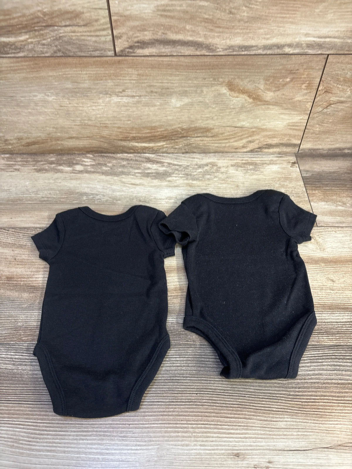Top view of two black Nike bodysuits in size 3 months showcasing short sleeves and rounded necklines on a wood-patterned floor