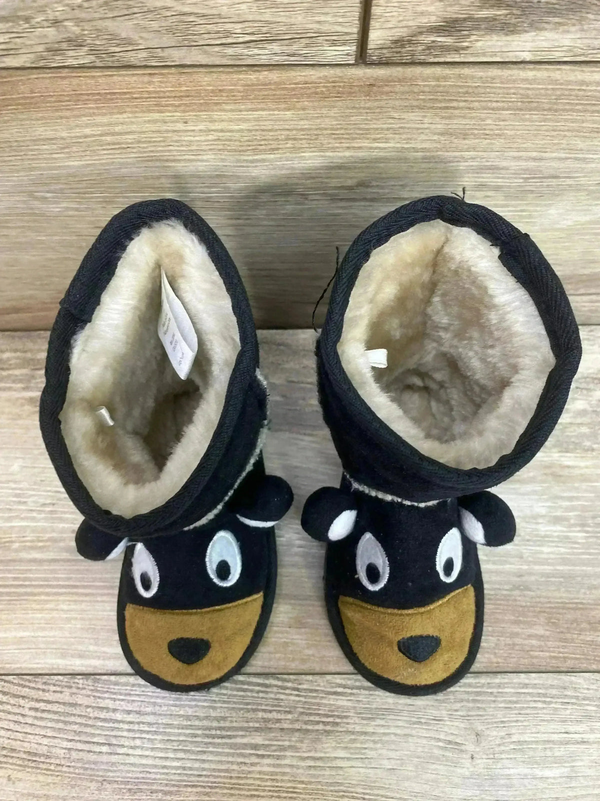 Top-down view of black kids’ moose slipper boots showing plush cream lining, embroidered moose face with ear details on wood
