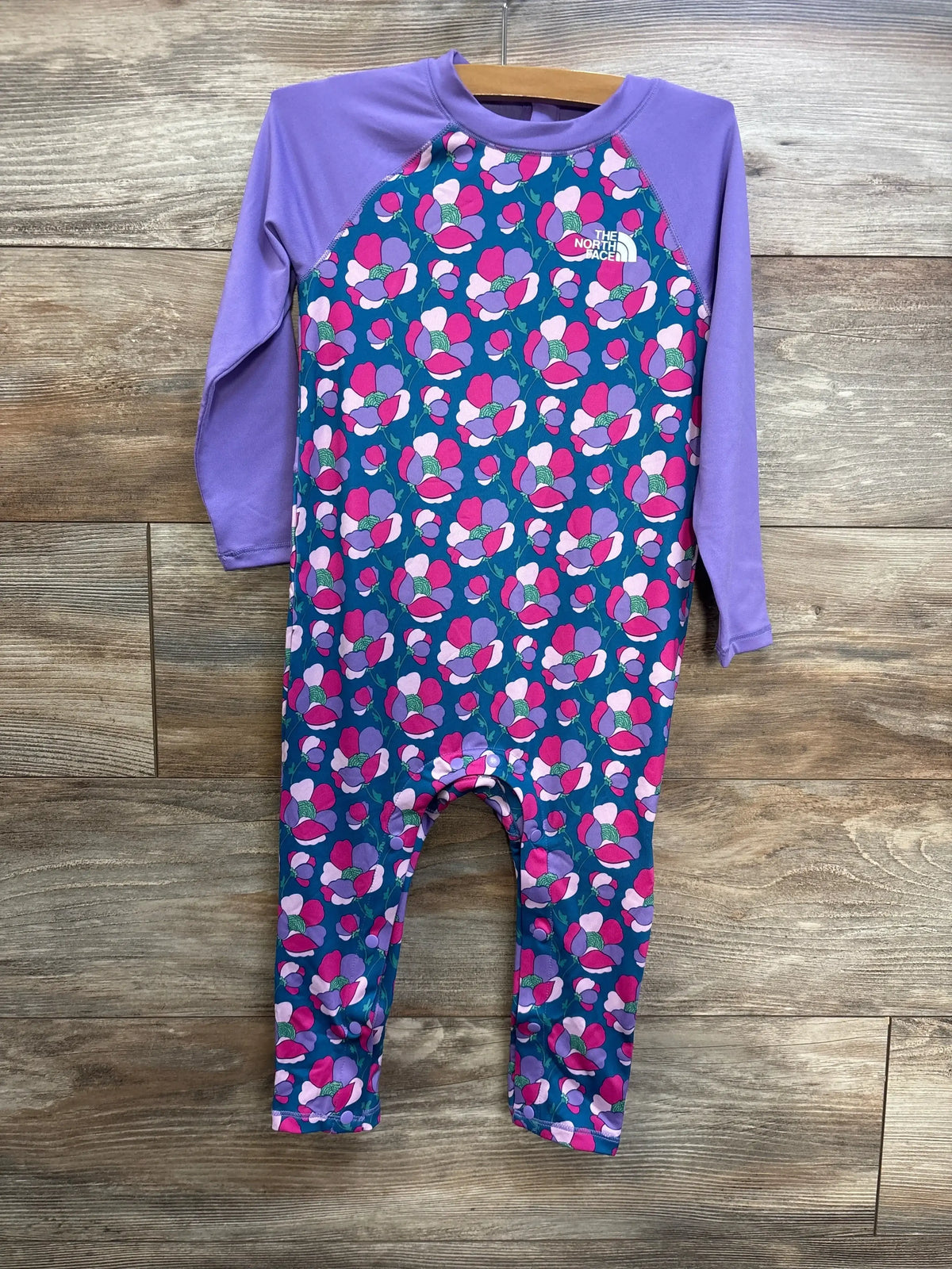 Front of The North Face baby sun rashguard coverall with purple sleeves, blue floral body and chest logo on wooden hanger