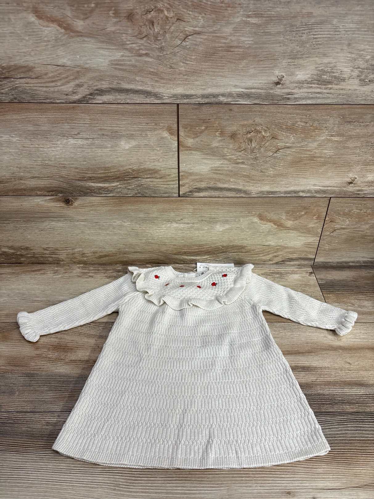White knitted long-sleeve baby dress with ruffled collar and red embroidered accents on wood background