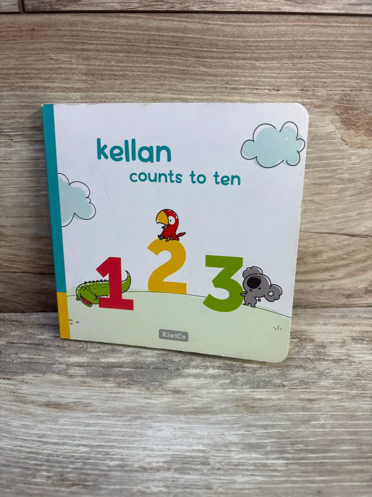 Front cover of KiwiCo Kellan Counts To Ten board book showcasing numbers 1, 2, and 3 with animal illustrations on a wood tabletop