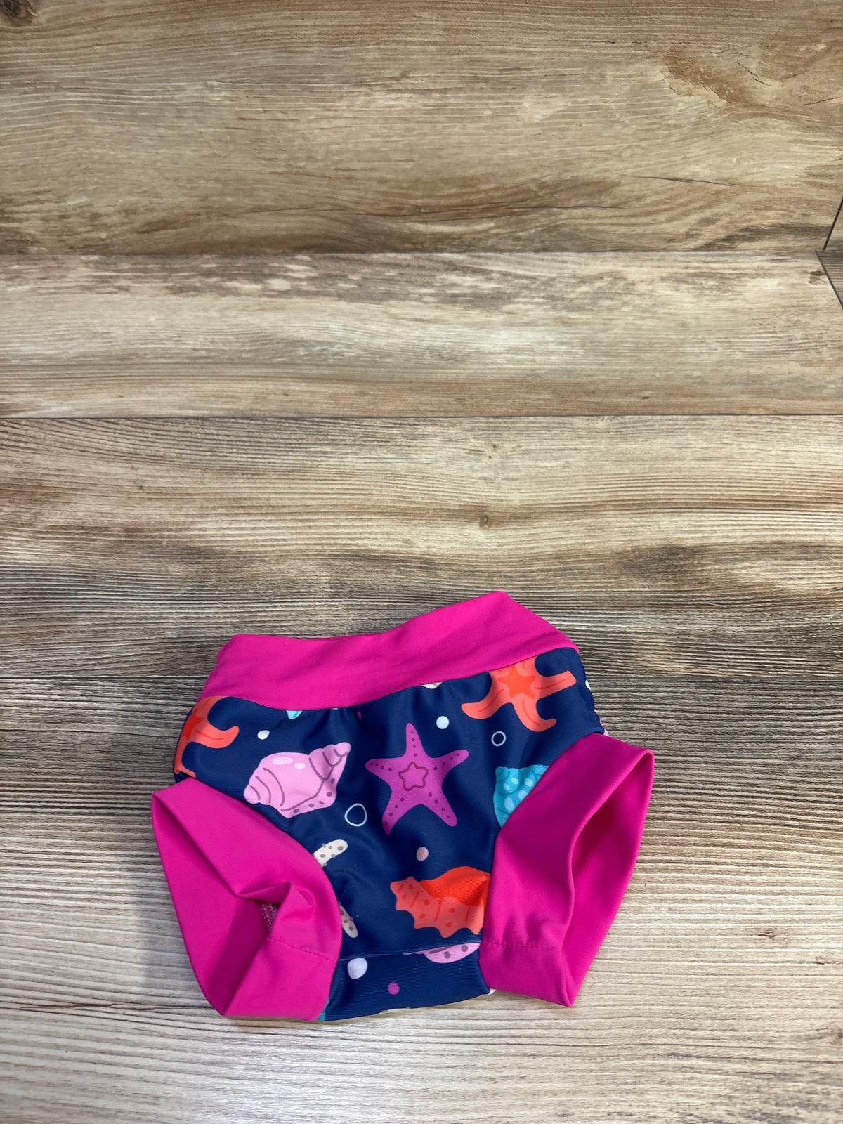 Flat lay of reusable swim nappy size 12 to 24 months showing navy sea creature print with pink waistband and leg cuffs on wood background