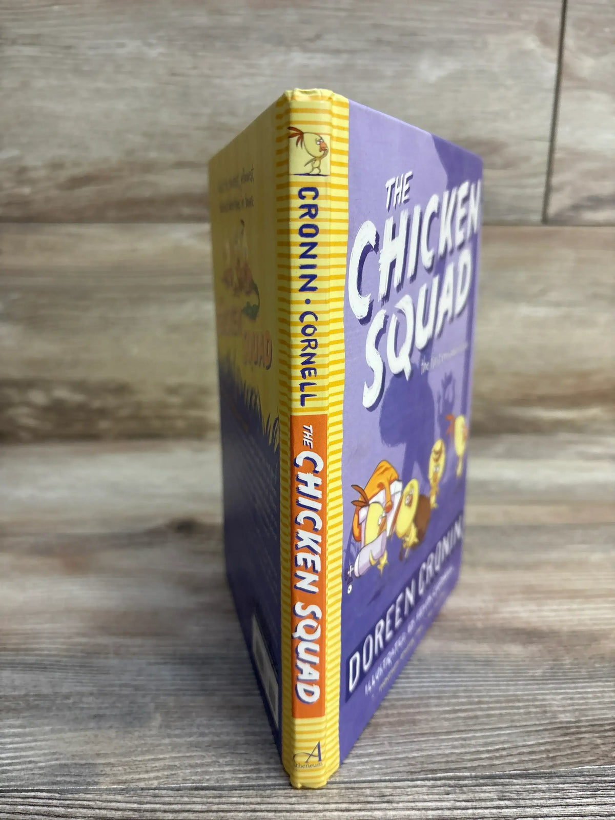 Angled view of The Chicken Squad hardcover book showing spine and partial front cover with purple illustrated cover on wood background