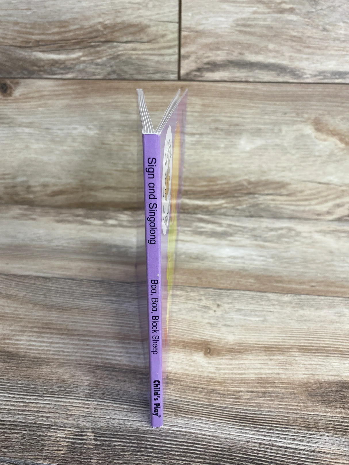 Side view of Sign and Sing Along: Baa Baa Black Sheep board book showing purple spine text and Child's Play logo on wood background