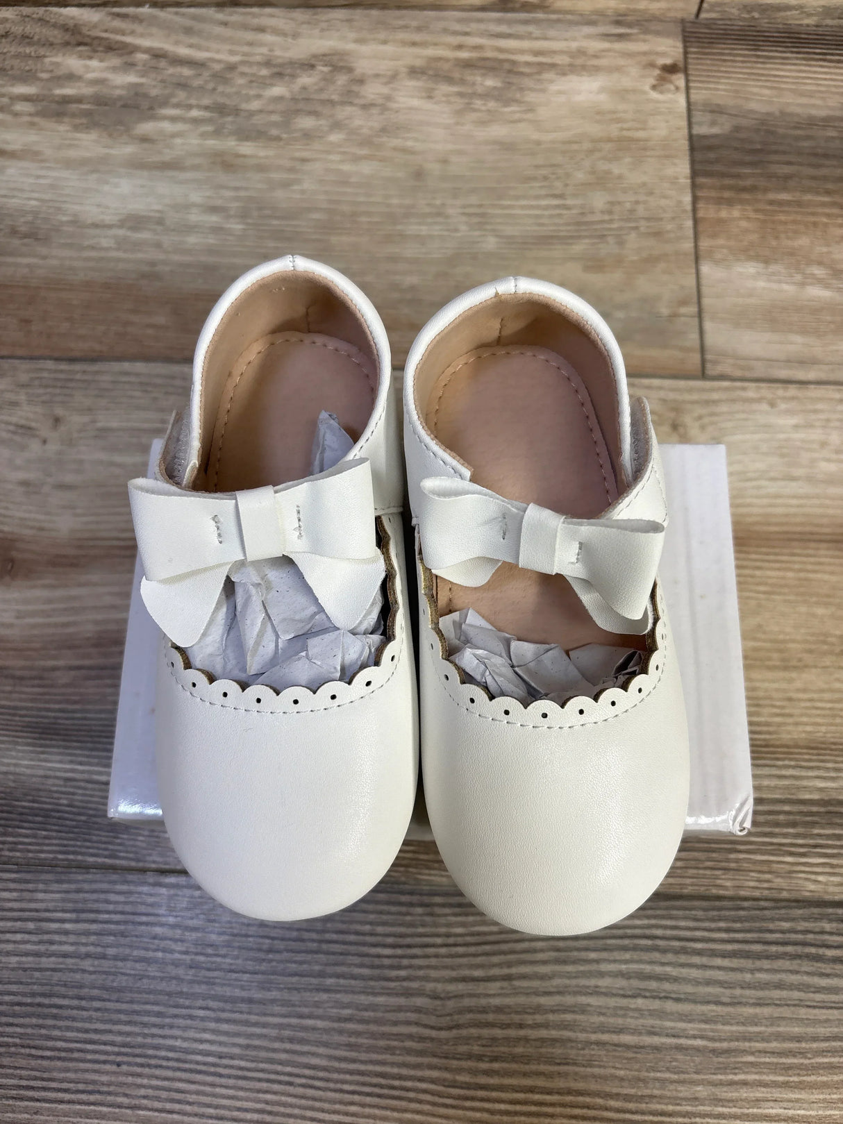 Top-down view of white leather baby princess flats size 7C showing bow straps and scalloped trim on a white box over wood floor