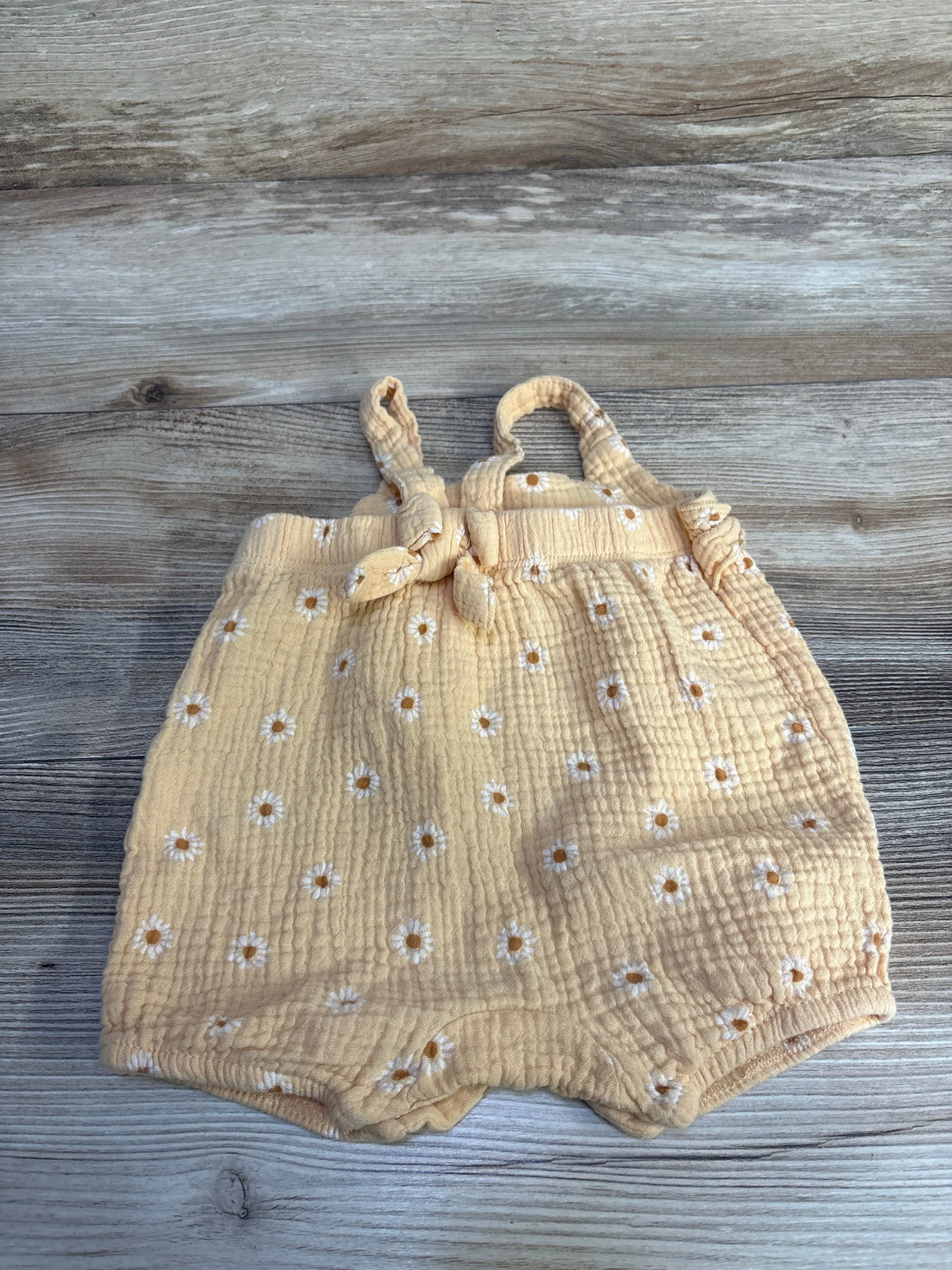 Front flat-lay of orange gauze baby romper with embroidered white daisies, knotted shoulder straps, and elastic leg openings on wood background