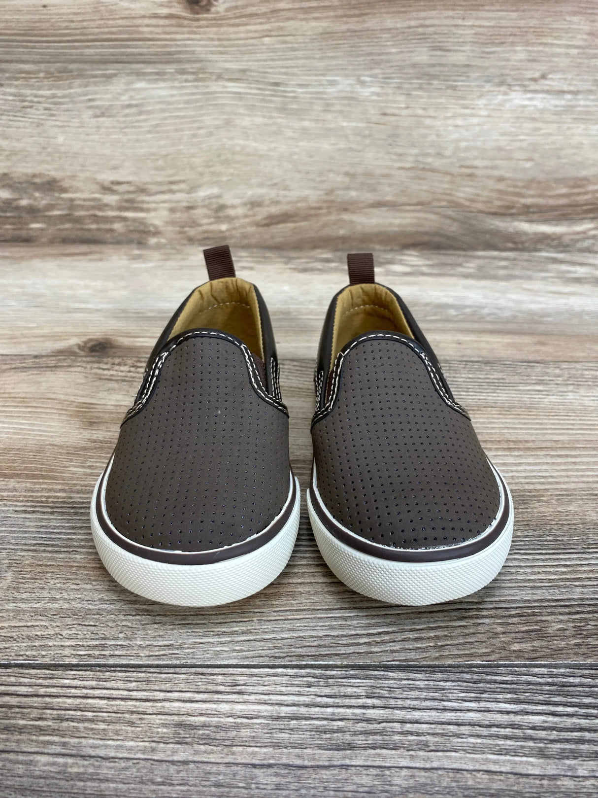 Front view of Gap toddler slip-on sneakers in brown showing perforated upper and white rubber sole on wood background