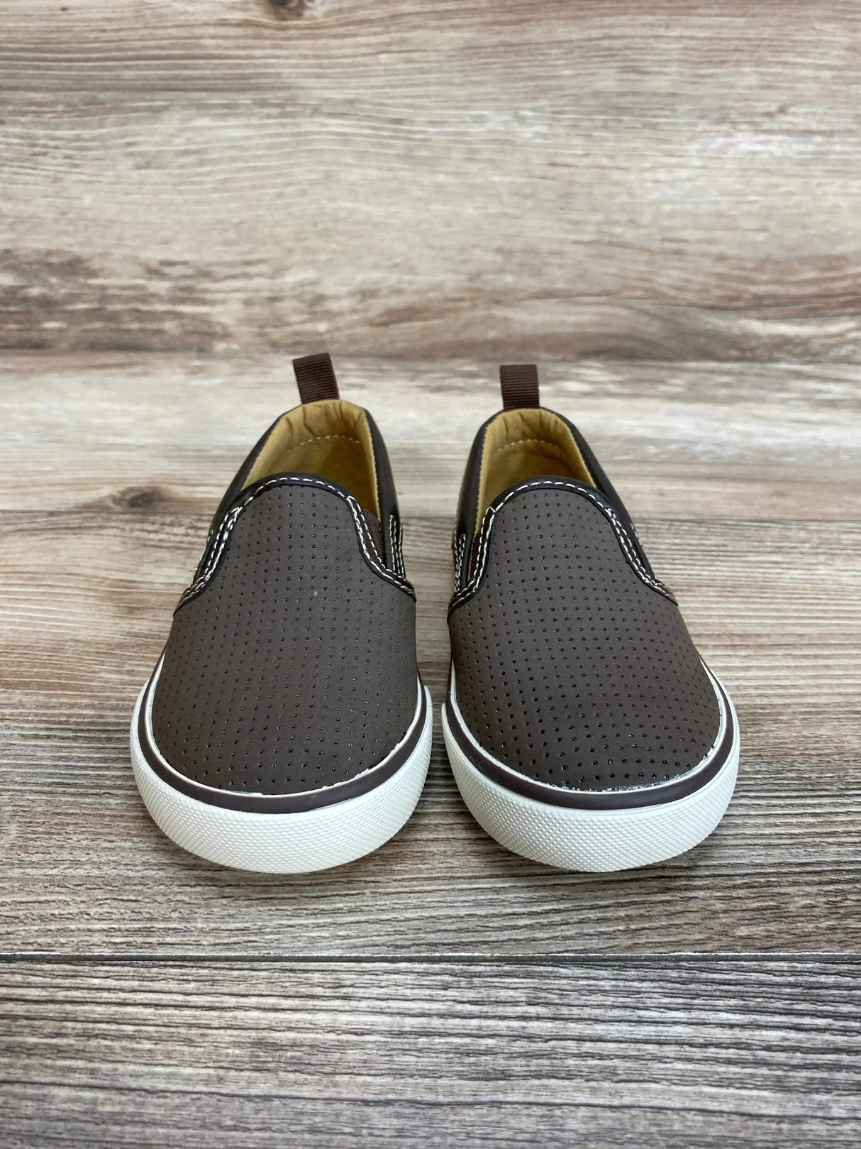 Front view of Gap toddler slip-on sneakers in brown showing perforated upper and white rubber sole on wood background