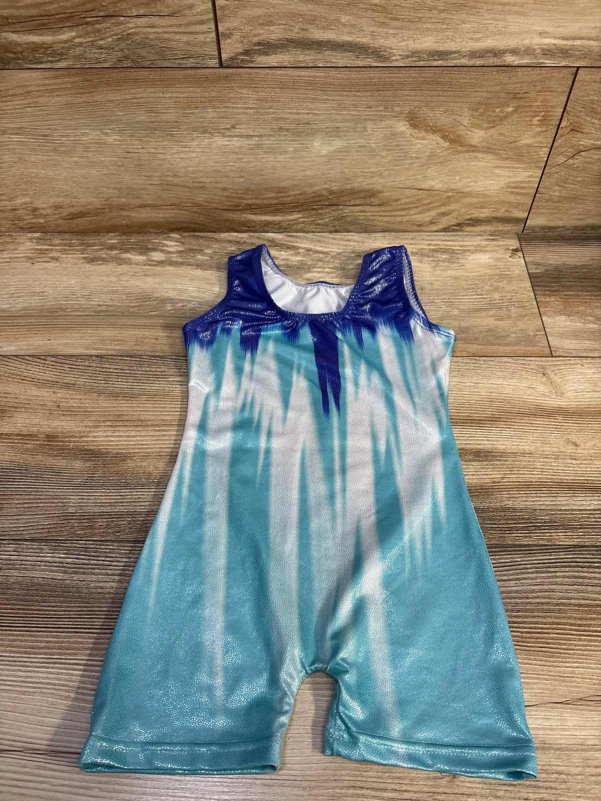 Flat lay of size 2/3T toddler gymnastics leotard showcasing teal-to-white ombre metallic fabric and shiny navy yoke on wood floor