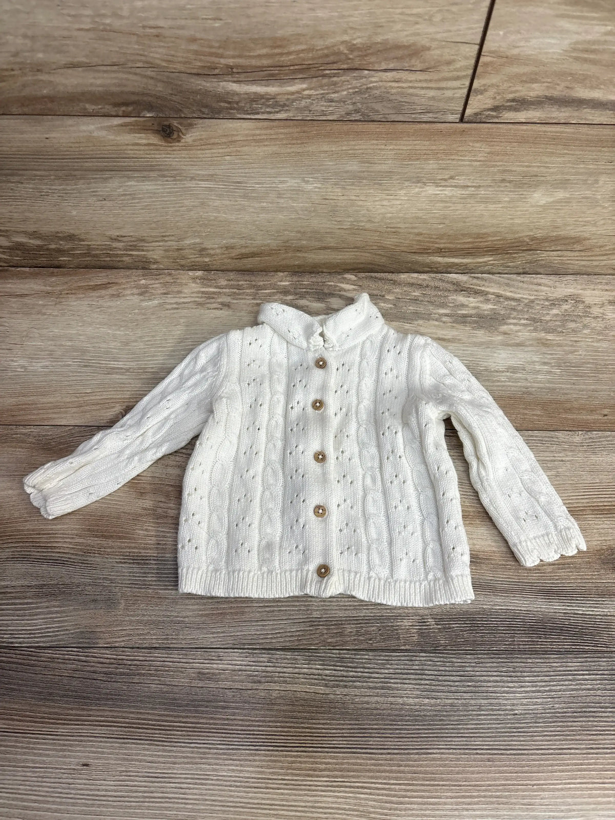 White knit baby cardigan sweater with buttons, long sleeves, cable knit pattern on wood background