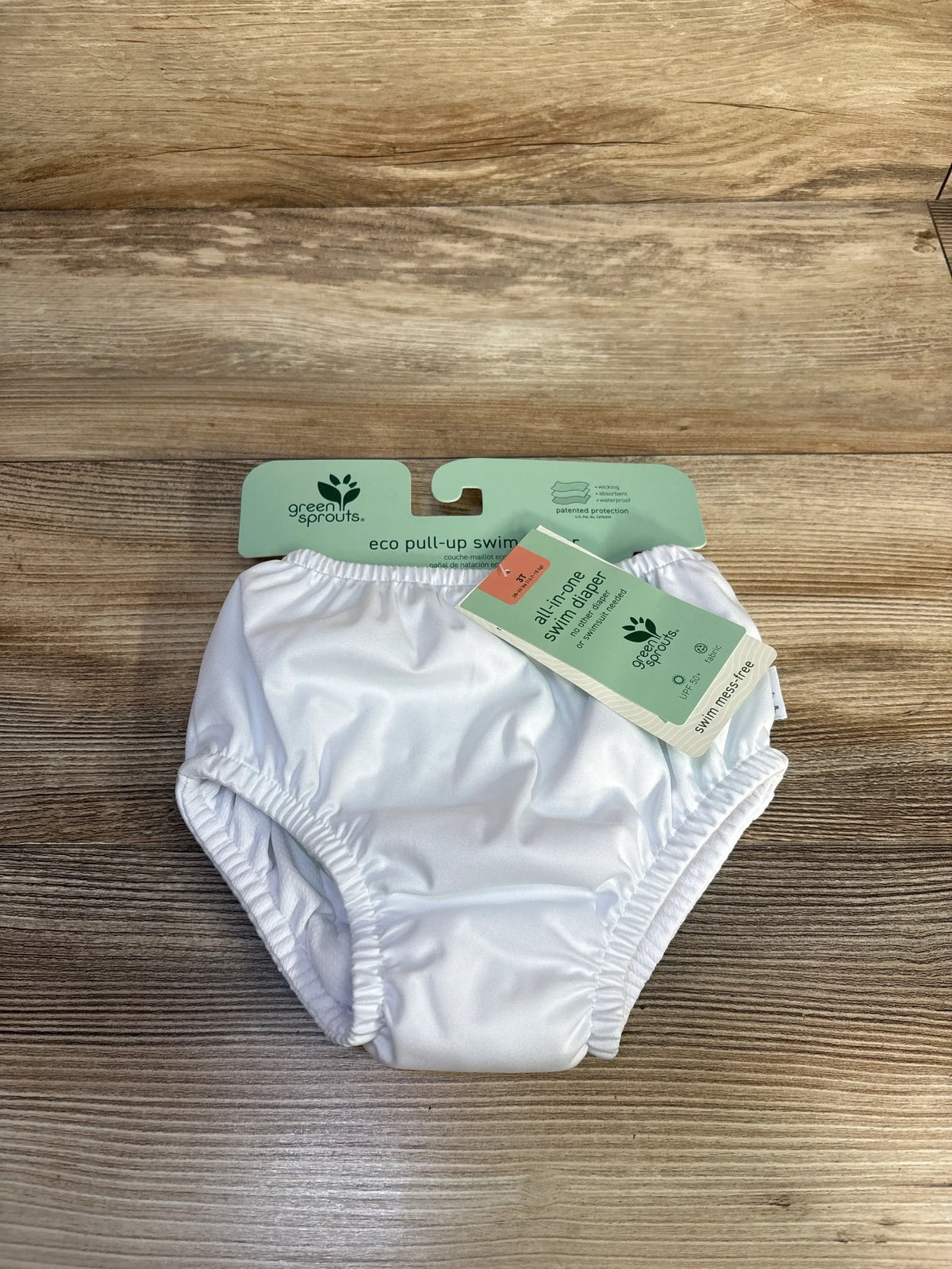 Front flat lay of Green Sprouts white eco pull-up swim diaper showing elastic leg openings and attached tag on wood