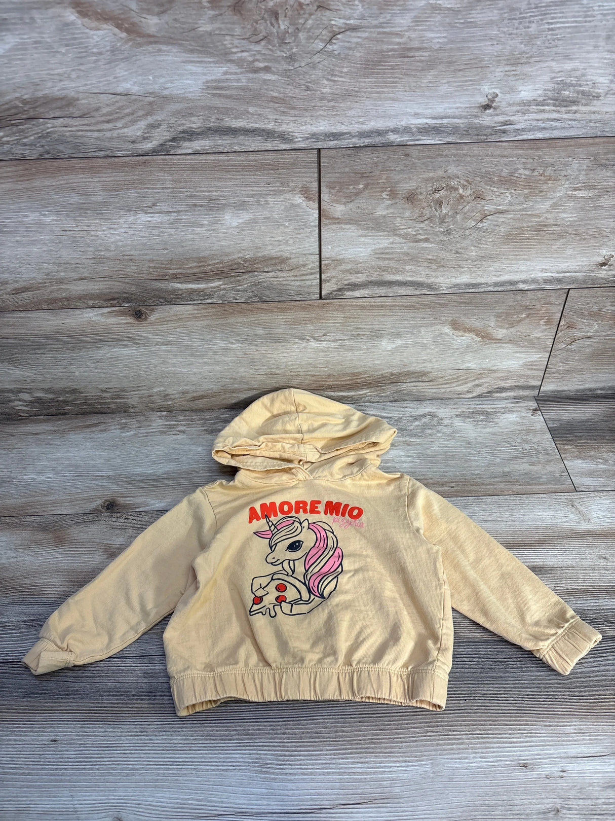 Beige kids hoodie with unicorn and pizza graphic, Amore Mio text, on wood background.