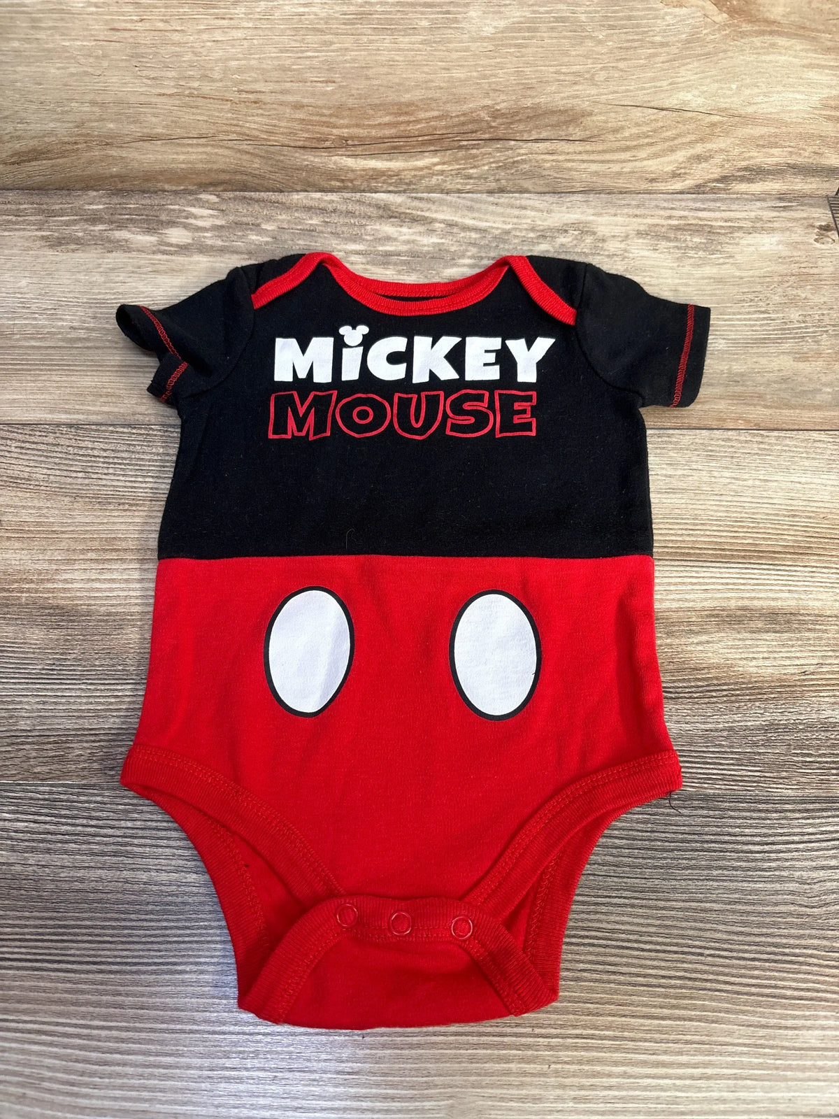 Front view of Disney Baby Mickey Mouse bodysuit showcasing black and red design with three snap closures on wood-grain background