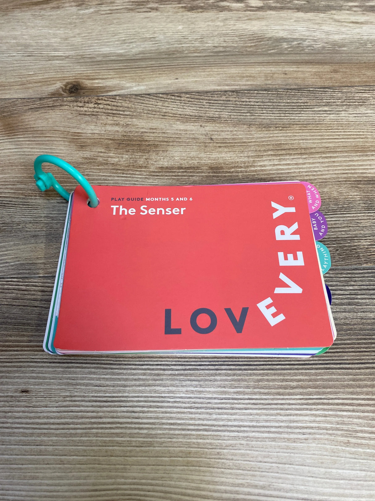 Top-down view of Lovevery The Senser play guide showing red cover, visible month tabs, and green ring binding on wooden surface