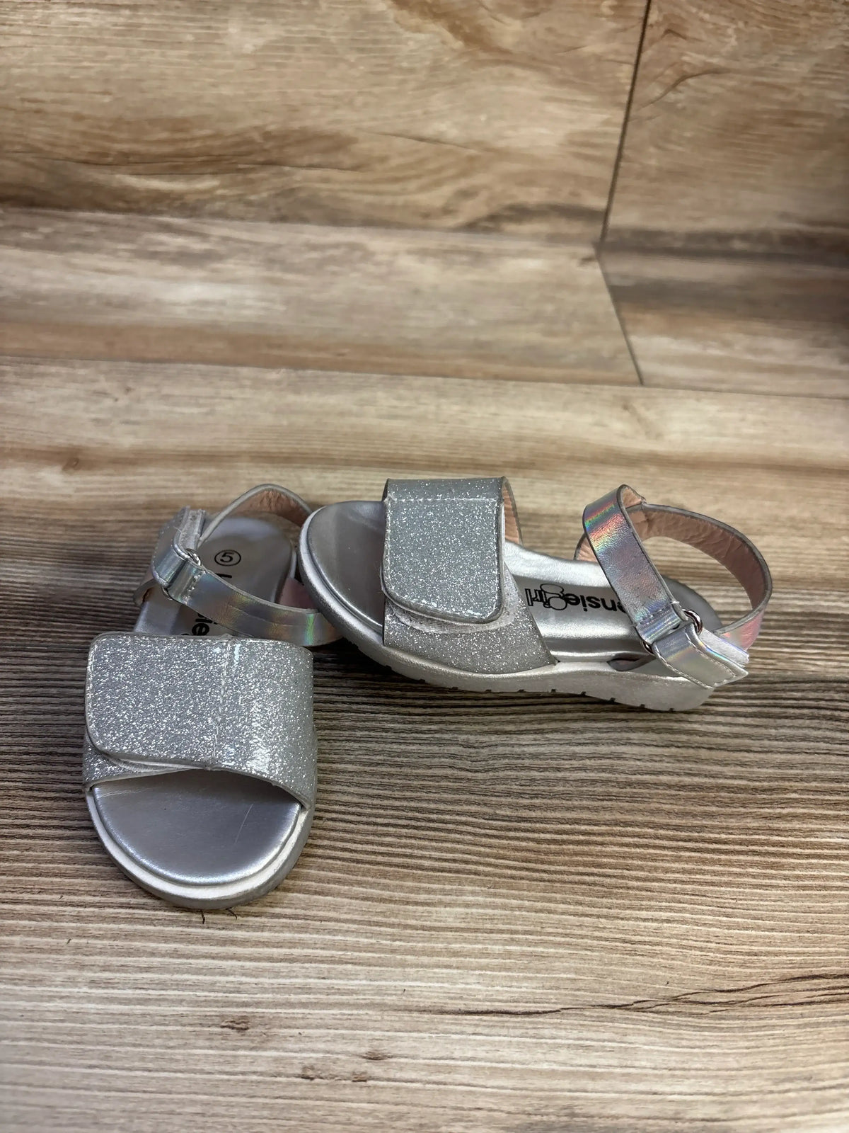 Front and angled view of Kensie Girl silver glitter sandals showcasing wide straps and ankle hook and loop closures on a wood look floor