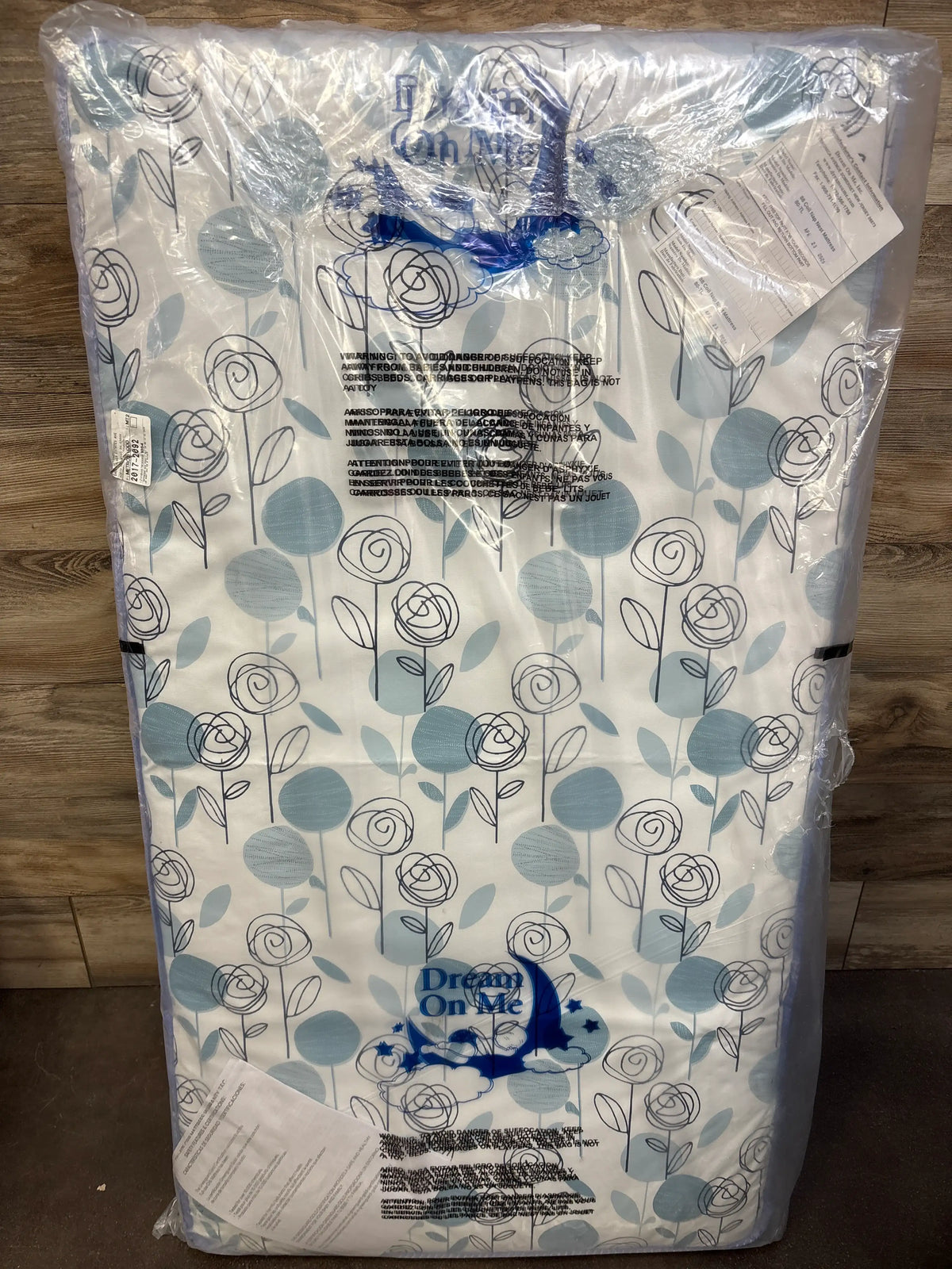 Front view of Dream On Me inner spring crib and toddler mattress showcasing a blue floral pattern in plastic packaging on a wood floor