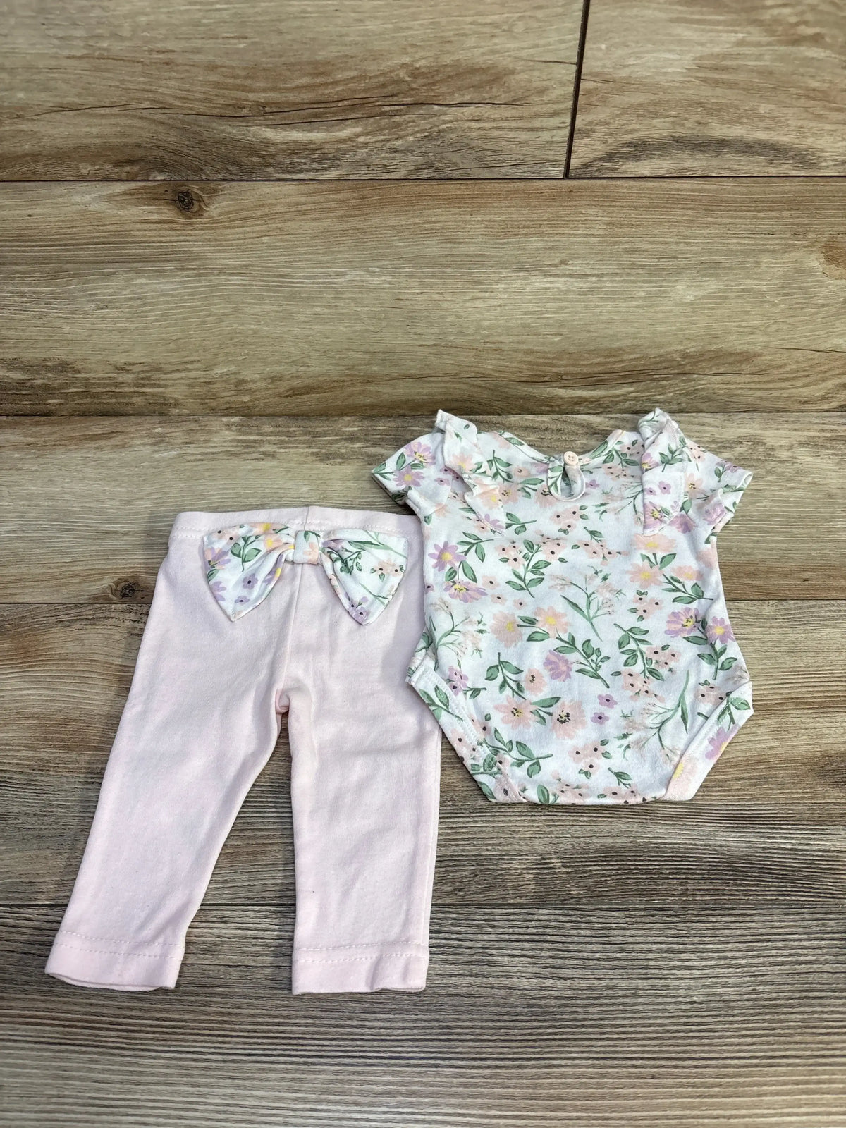 Flat lay of 2-piece baby set: floral short-sleeve bodysuit and pink pants with matching floral bow on wood background