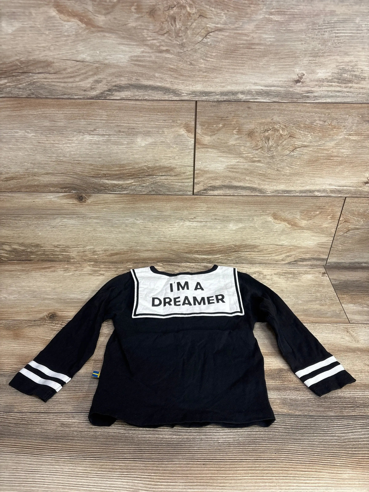 Front view of a black long sleeve toddler shirt showcasing sleeve stripes, a white collar, and I’M A DREAMER text on a wood floor