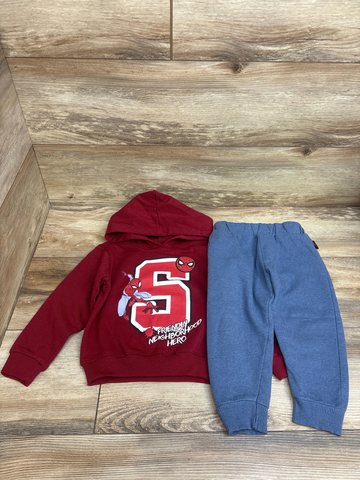 Top view of Marvel Spider-Man two-piece set in size 3T, with a red graphic hoodie and blue joggers on a wood-look floor
