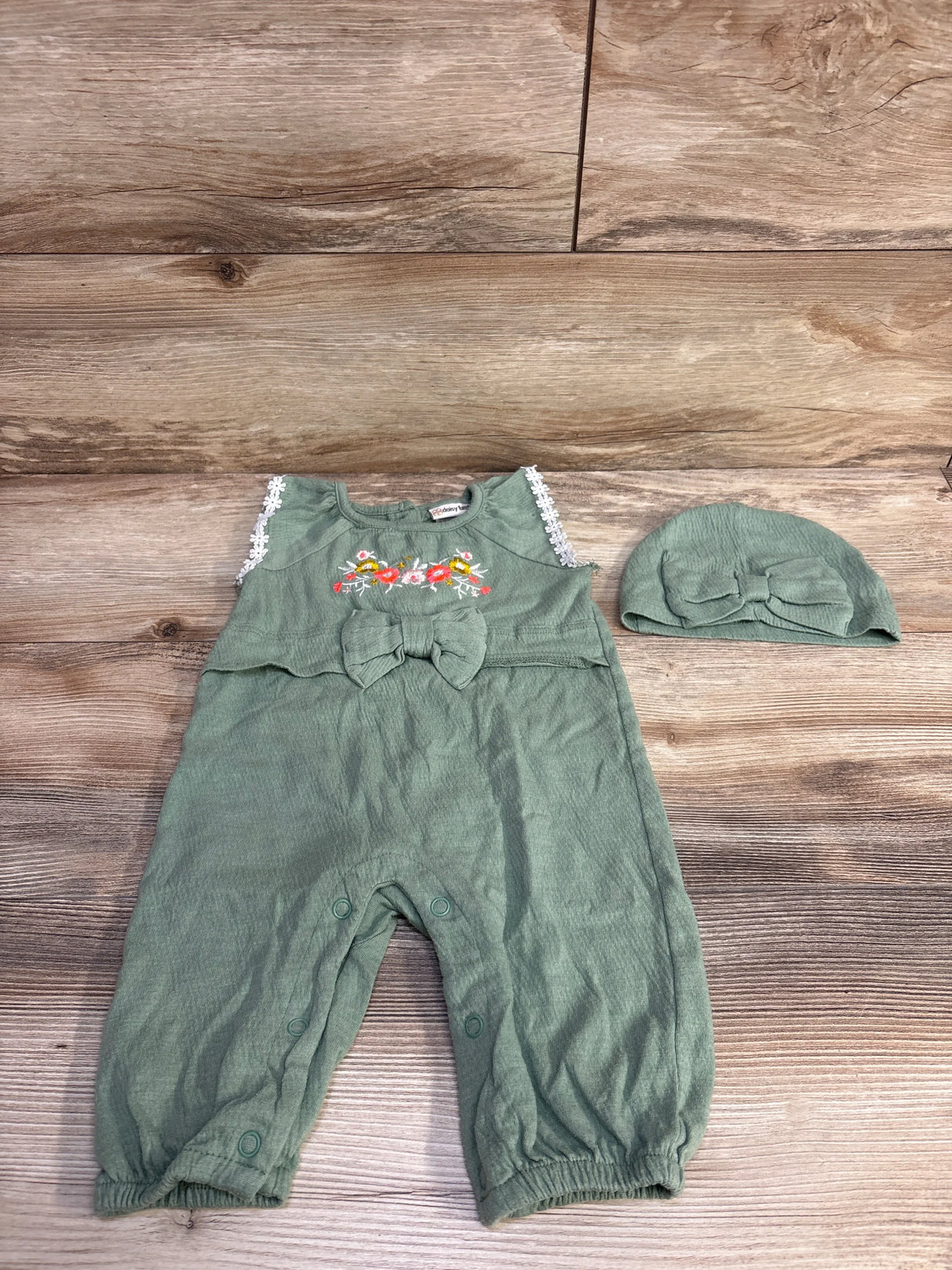 Front flat lay of green baby romper and matching hat showing floral embroidery, bow and lace trim on wood background