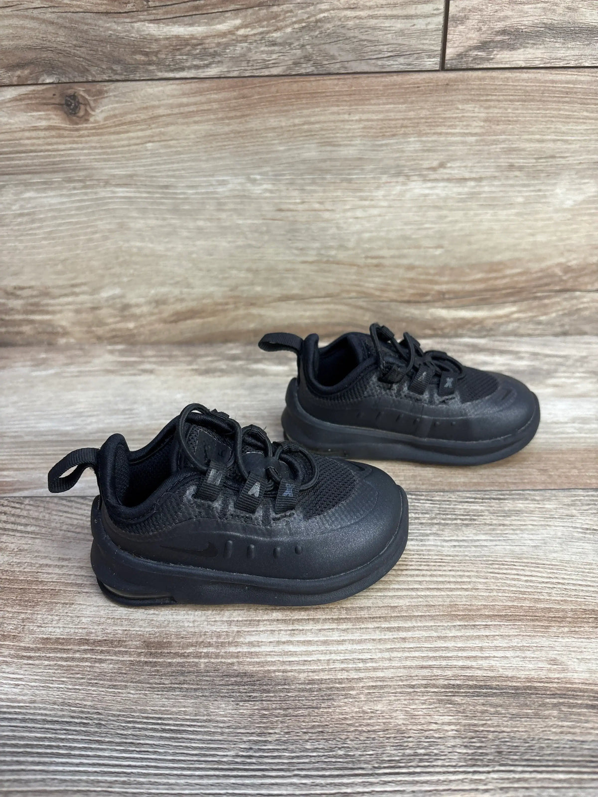 Angled view of Nike Air Max Axis black toddler sneakers with mesh upper, pull tab and cushioned sole on wood background